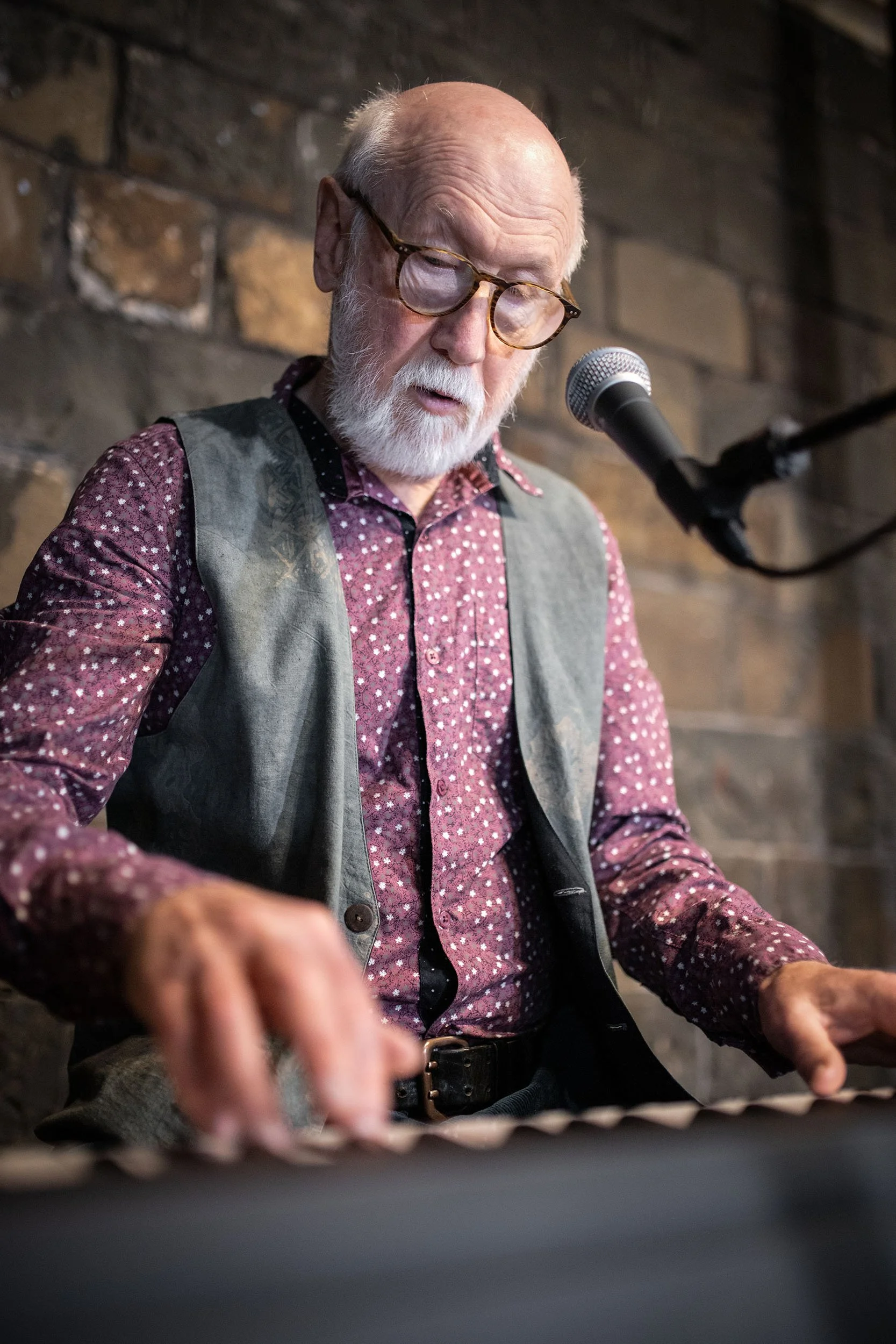 Alan Reid, a master storyteller in song and former member of Battlefield Band, playing a keyboard at a music venue with a brick wall in the background.