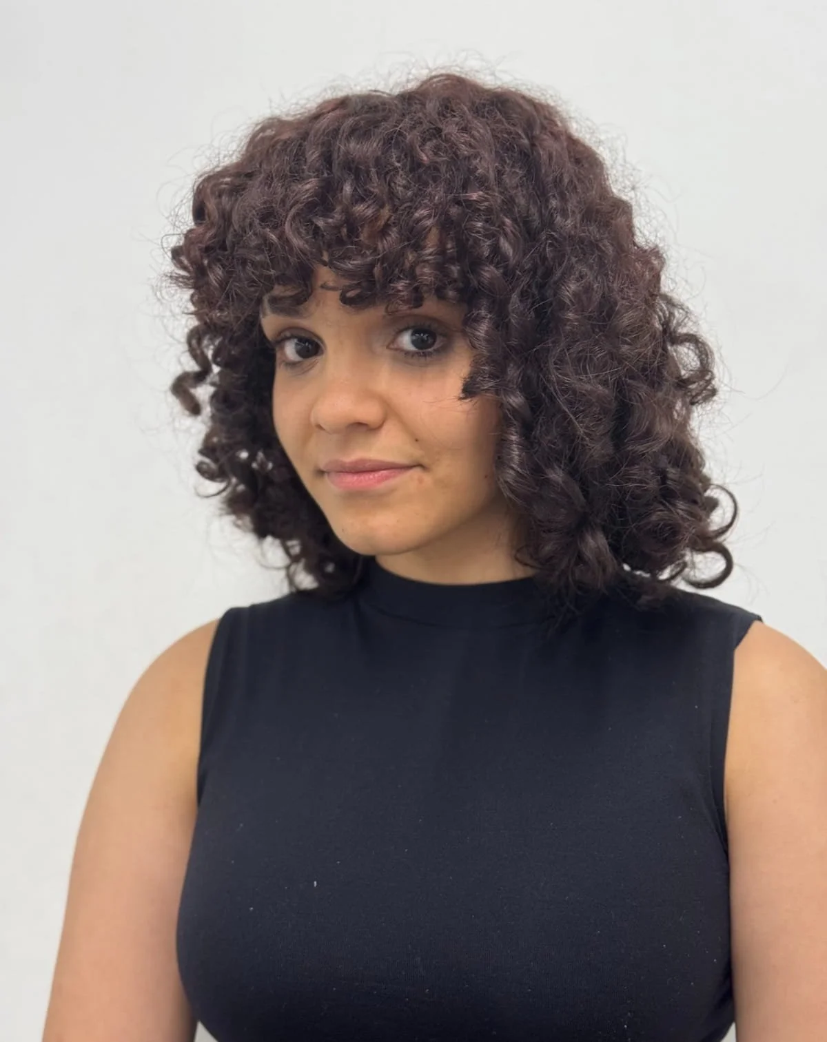 Portrait of actor Summer Currie, with a curly fringe wearing a black sleeveless top. Shot against white background.