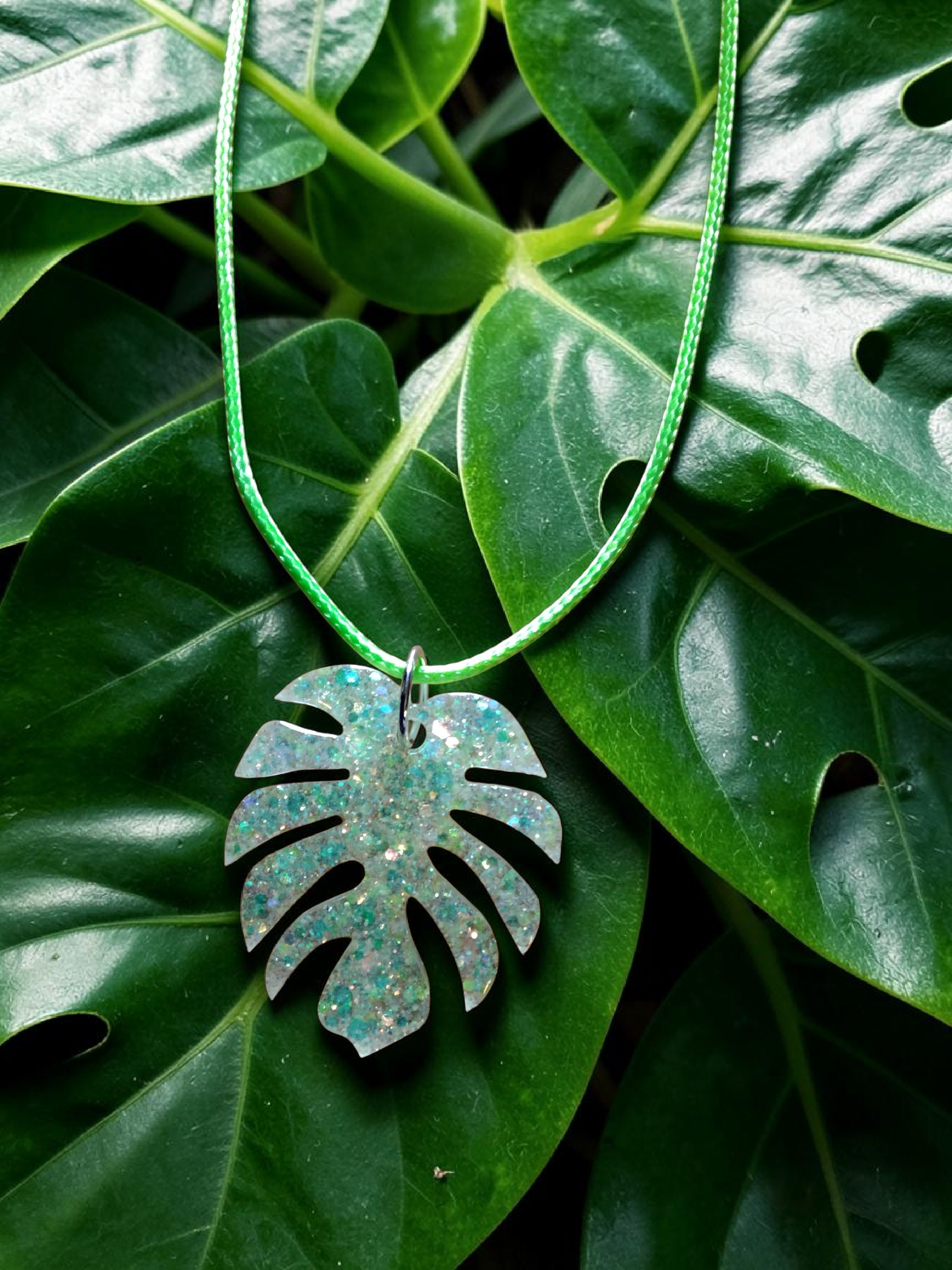 A decorative necklace with a green and white braided cord and a large, glittery monstera leaf-shaped pendant, placed on dark green tropical leaves.