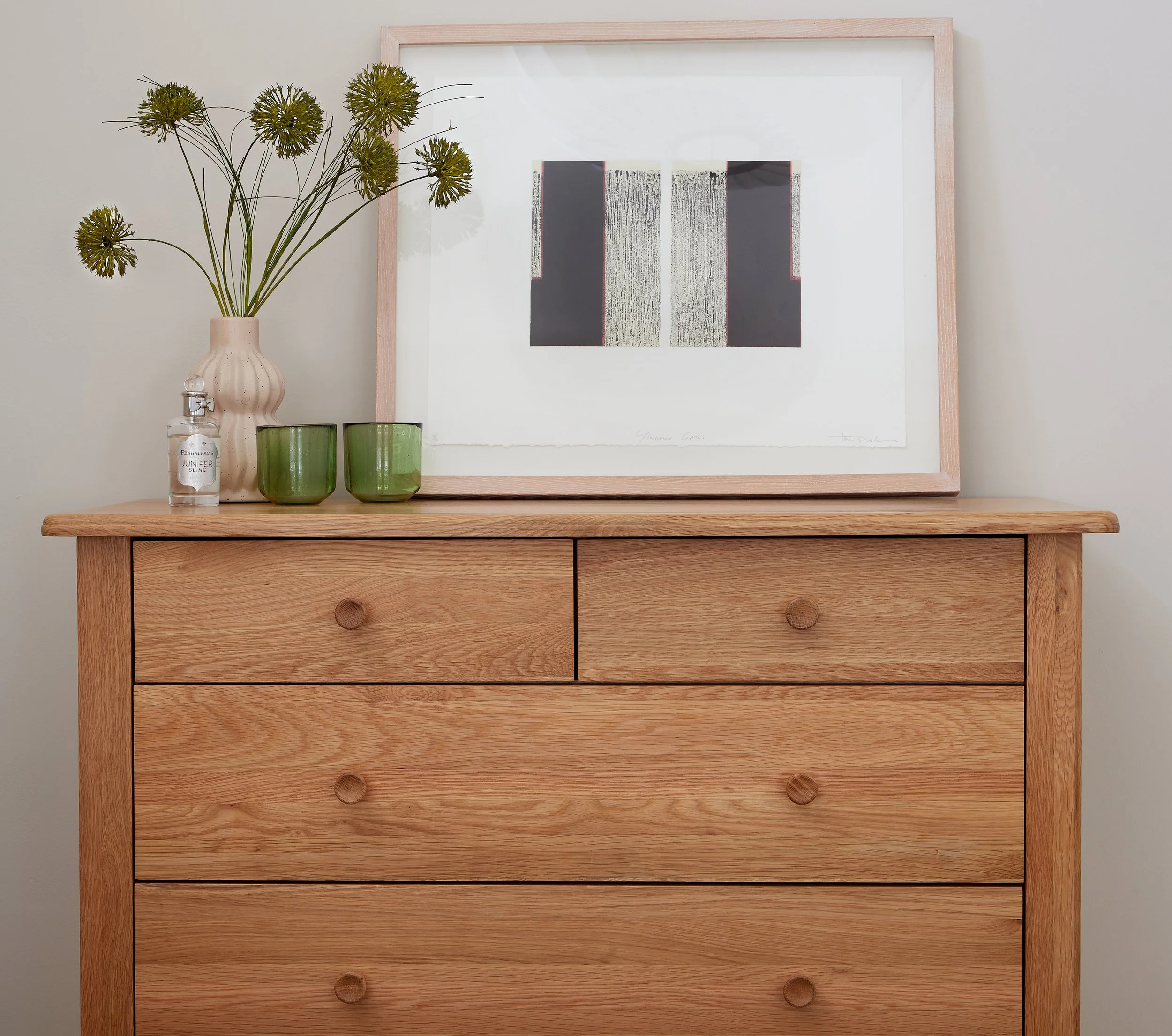 A wooden dresser with an abstract art print, two green cups, a clear hand sanitizer bottle, and a beige vase with green flowers.