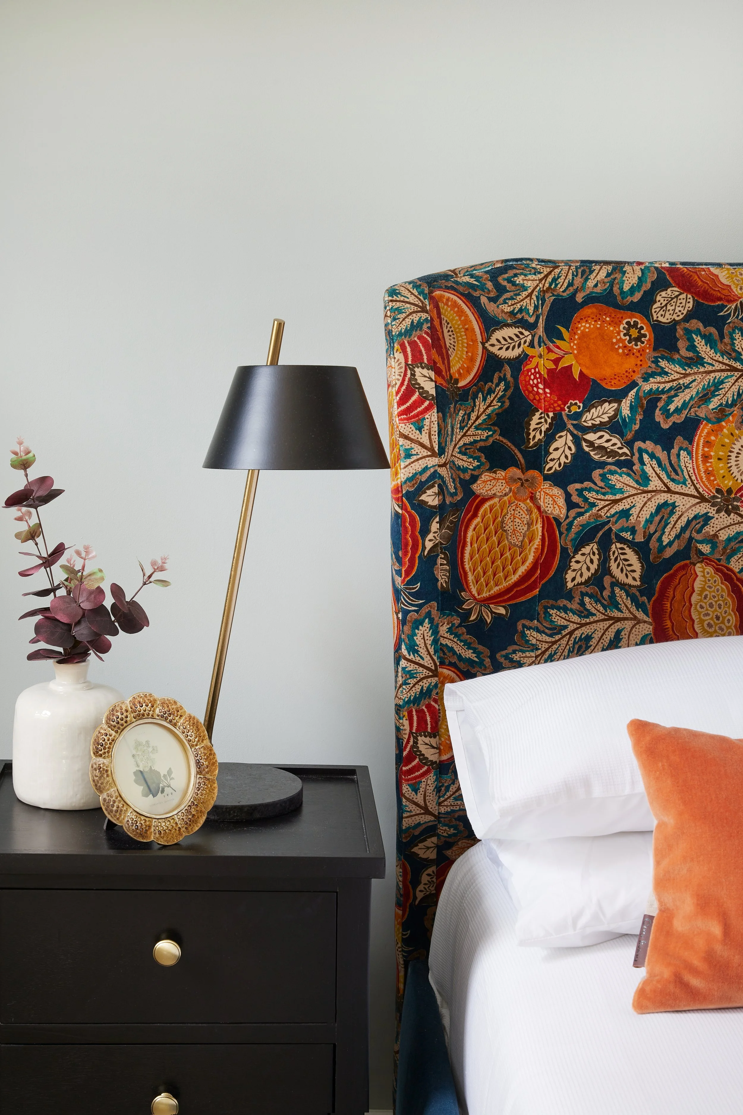 A black nightstand with a lamp, a vase with purple leaves, and a framed flower picture, next to a bed with a colorful, floral-patterned headboard and a white pillow with an orange pillow.