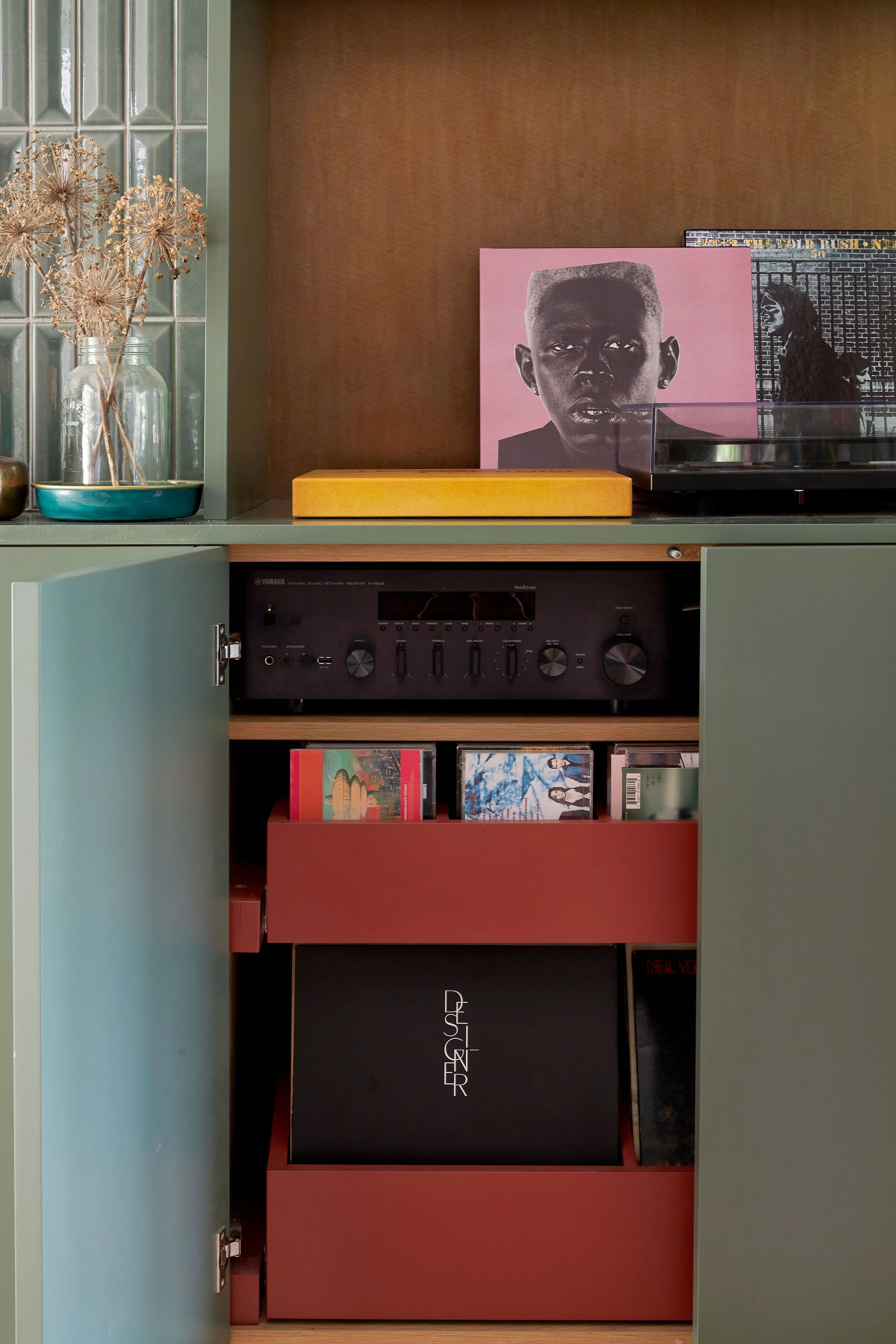 A wooden cabinet with artwork and vinyl records inside, a turntable on top, and a dried flower arrangement in glass jars on the left.