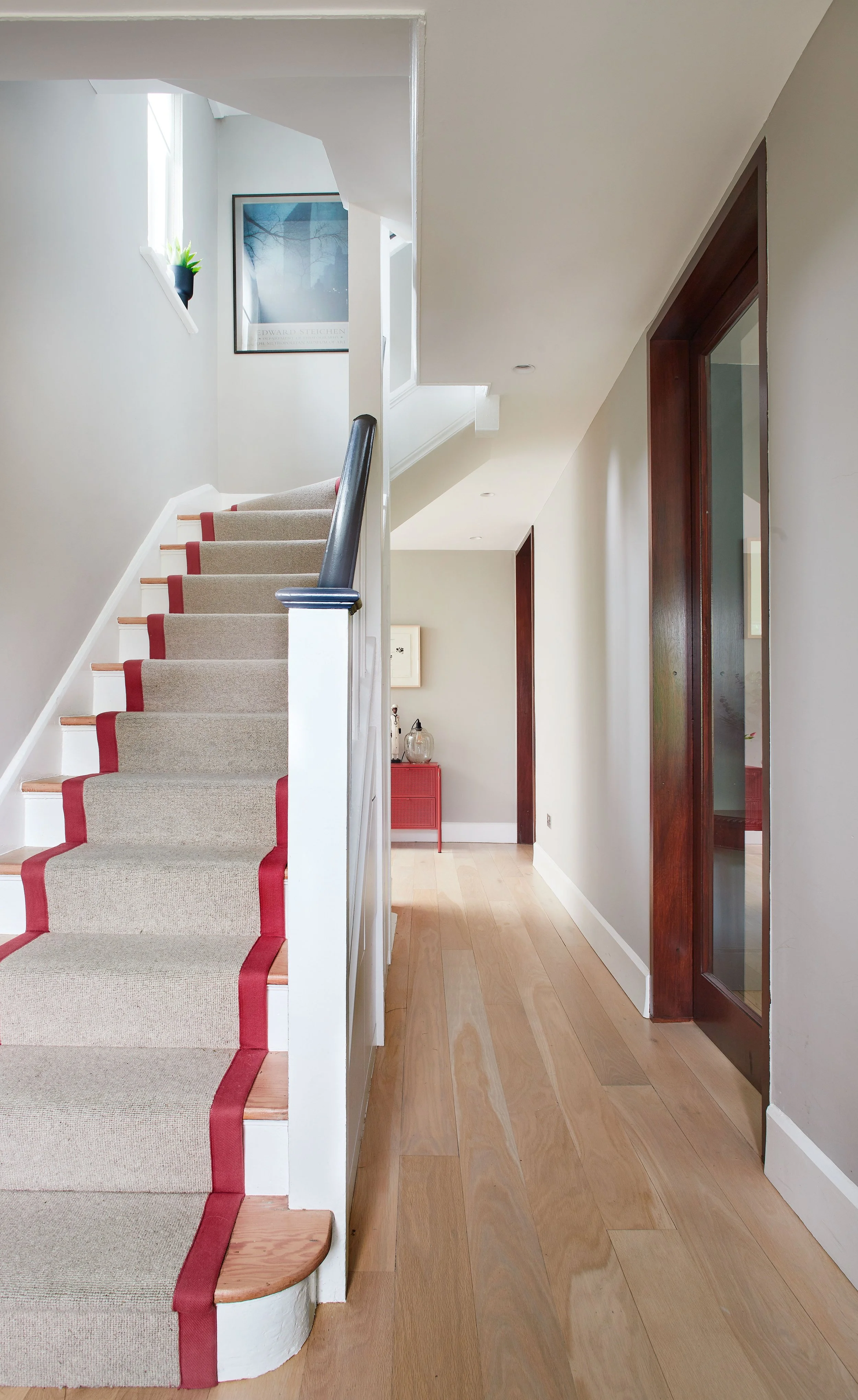 Interior view of a house with a staircase, light wood flooring, and white walls. The staircase has a beige carpet runner with red accents and a black handrail. There are framed artworks on the walls, a small red table with decorative items, and a pot