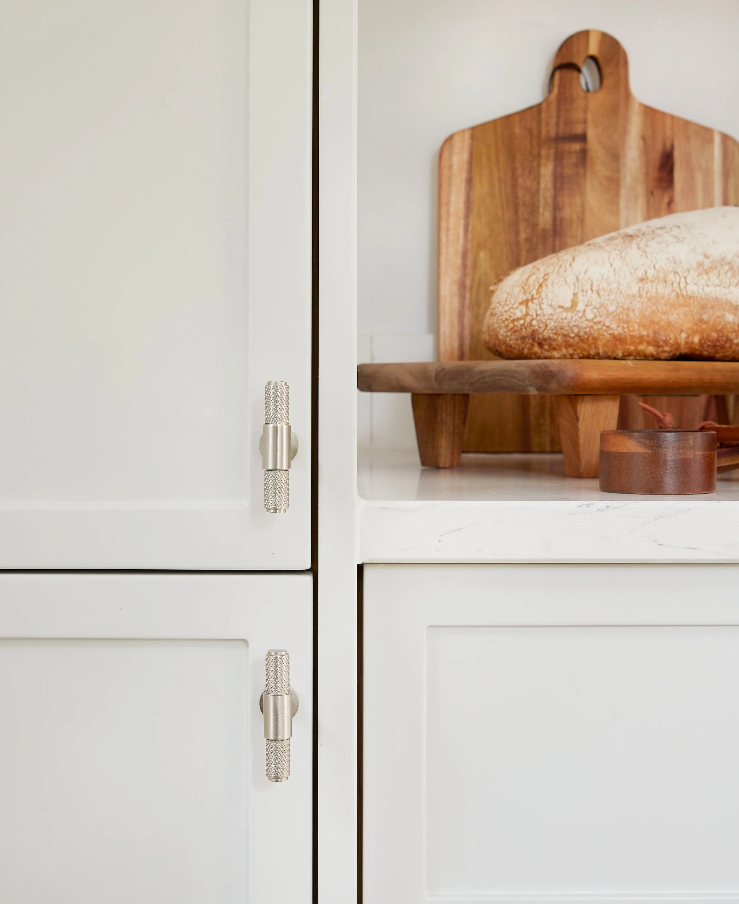 Close-up of a white kitchen cabinet door with metal hinges, next to a cut-out shelf with a wooden cutting board and a loaf of bread.