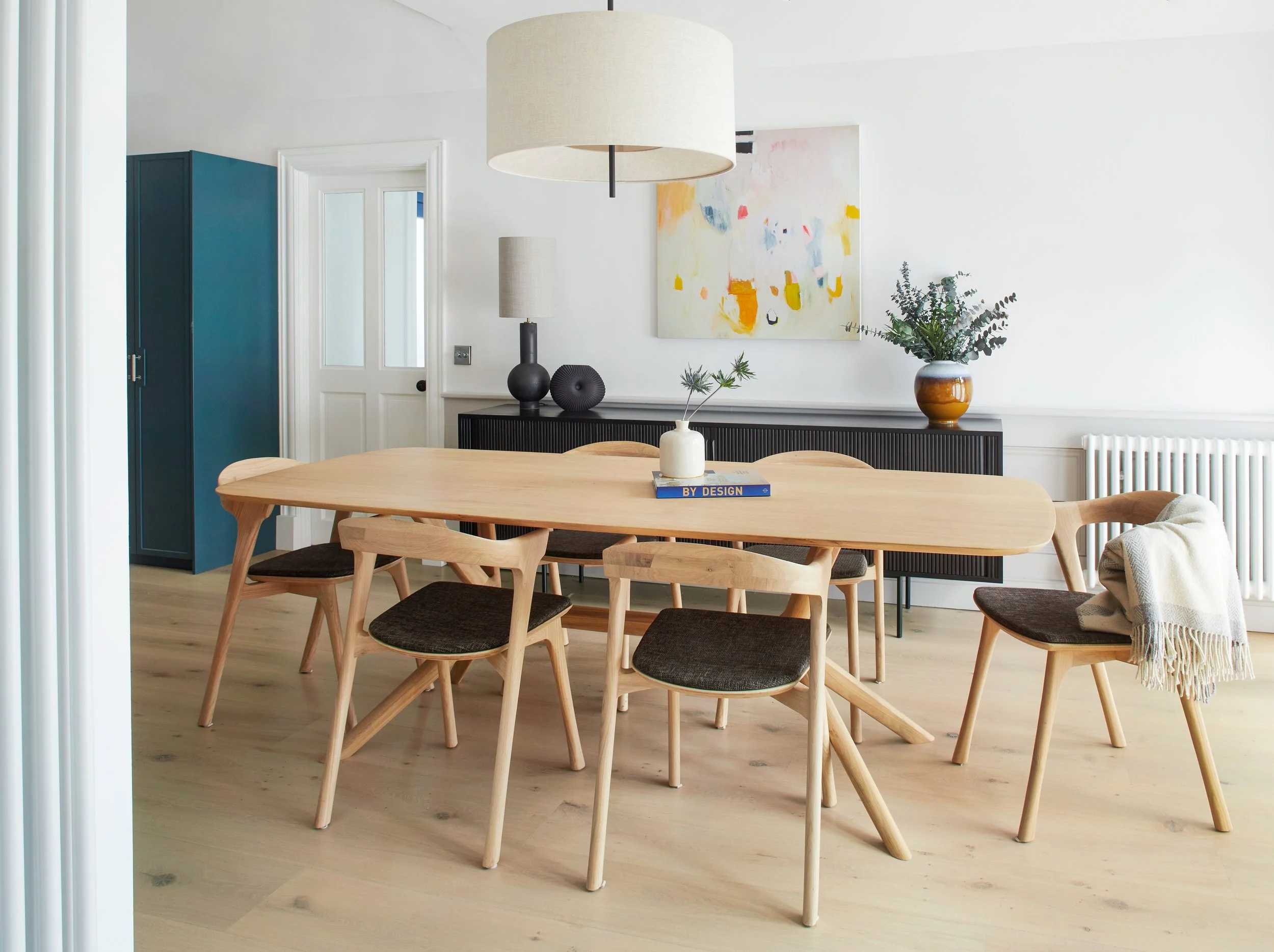 Modern dining room with wooden table and six chairs, minimalist decor, black sideboard, abstract wall art, and houseplants.
