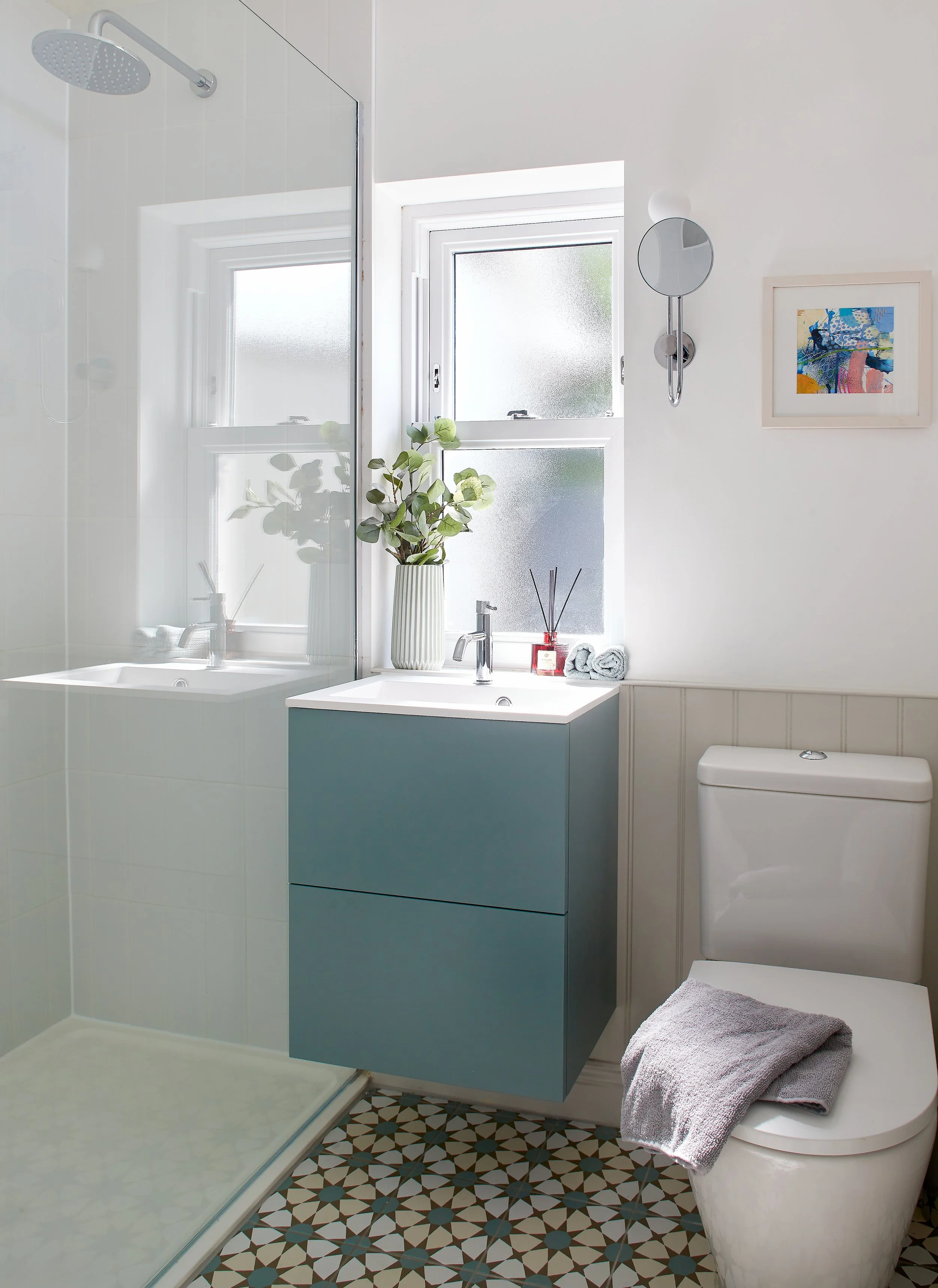 A modern bathroom with a walk-in shower, a blue vanity with a white sink, a frosted window with a plant, a framed artwork, a white toilet with a towel, and patterned tile flooring.
