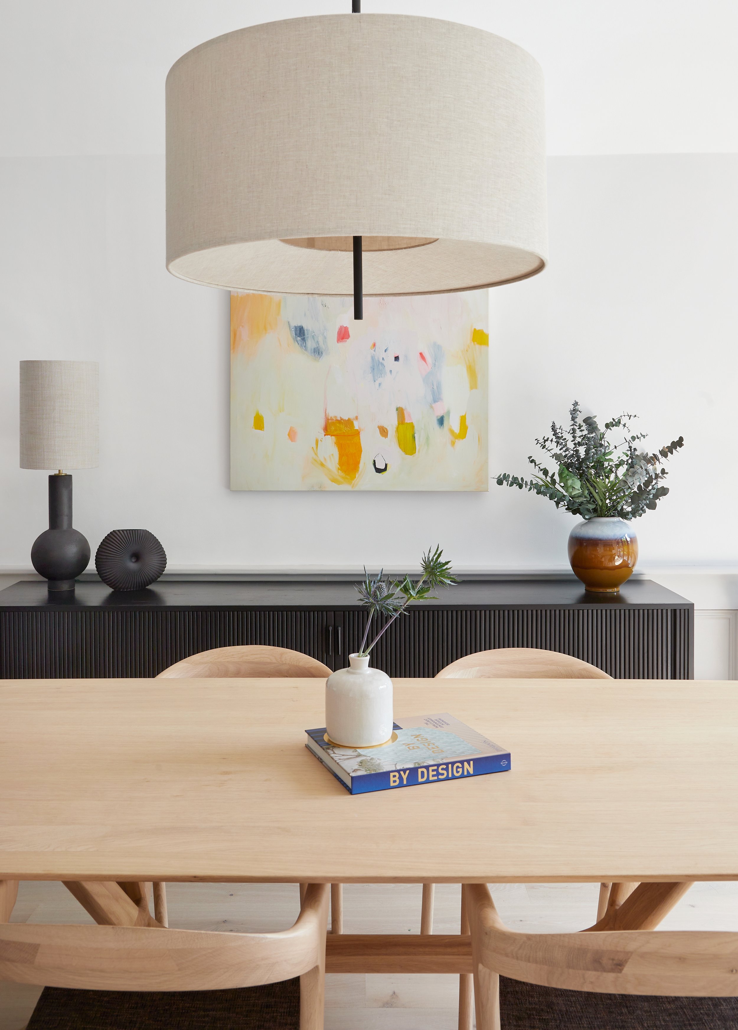 Dining room with light wooden table, beige chairs, large beige pendant lamp, black sideboard, colorful abstract painting, decorative vases, and a small plant on the table.