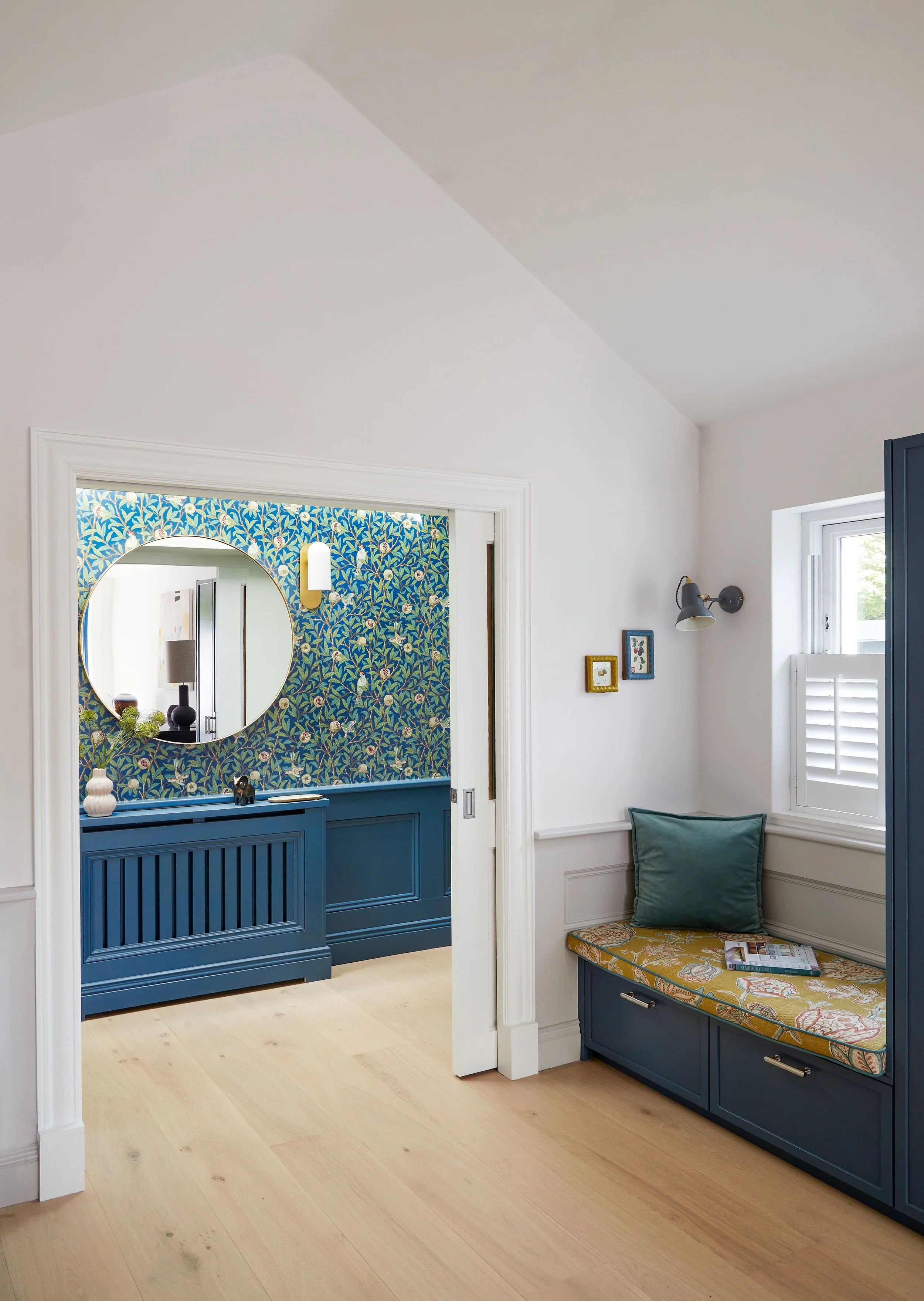 A bright room with white walls and light wood flooring. On the right side, there is a window with white shutters, and a built-in bench with a cushion and some magazines. On the left, a doorway with a decorative white frame leads to a room with blue w