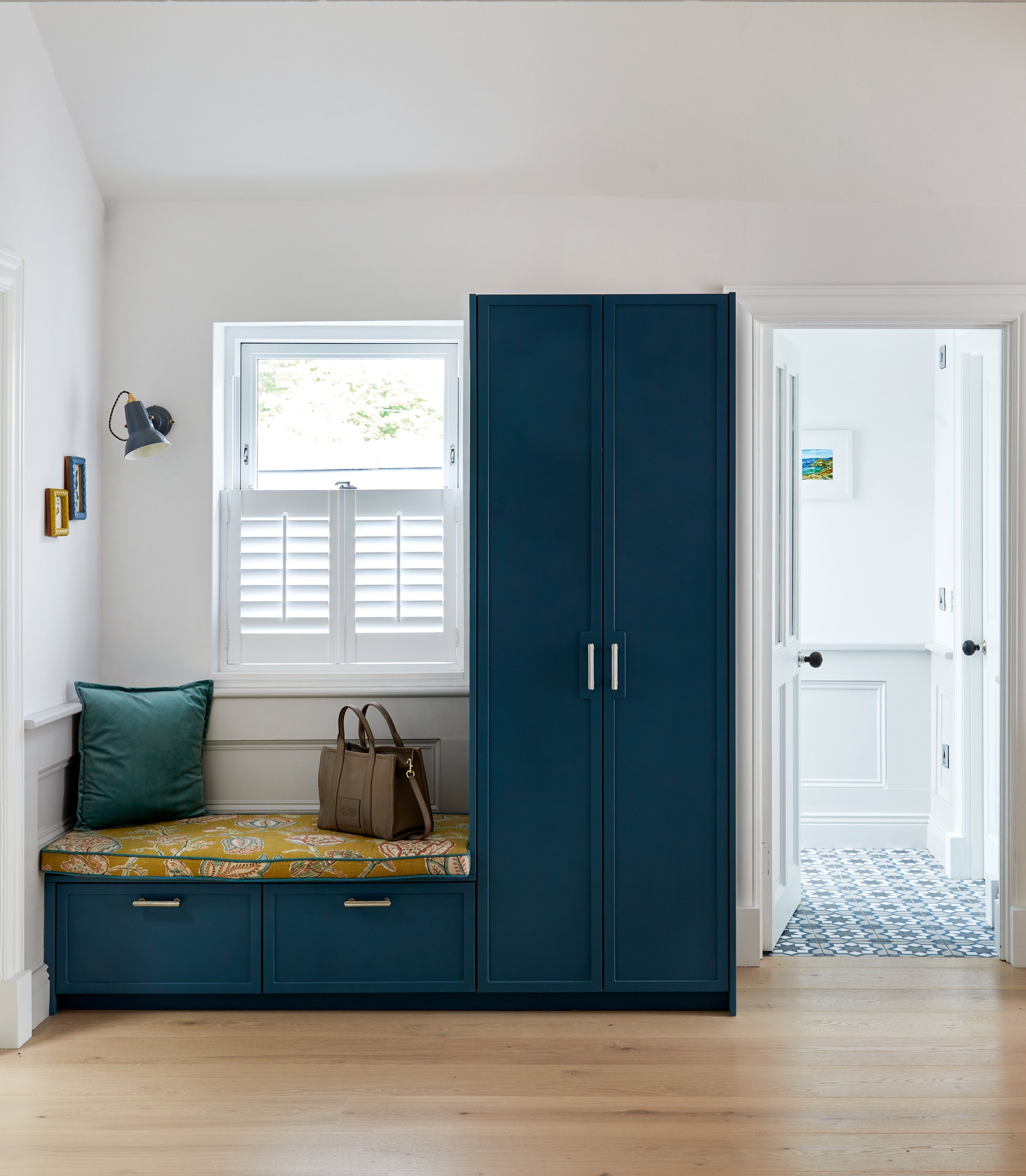 A cozy entryway with a built-in bench and blue cabinet, a window with white shutters, a small brown handbag on the bench, and a wall-mounted lamp, leading to a bathroom with patterned tiled floor.
