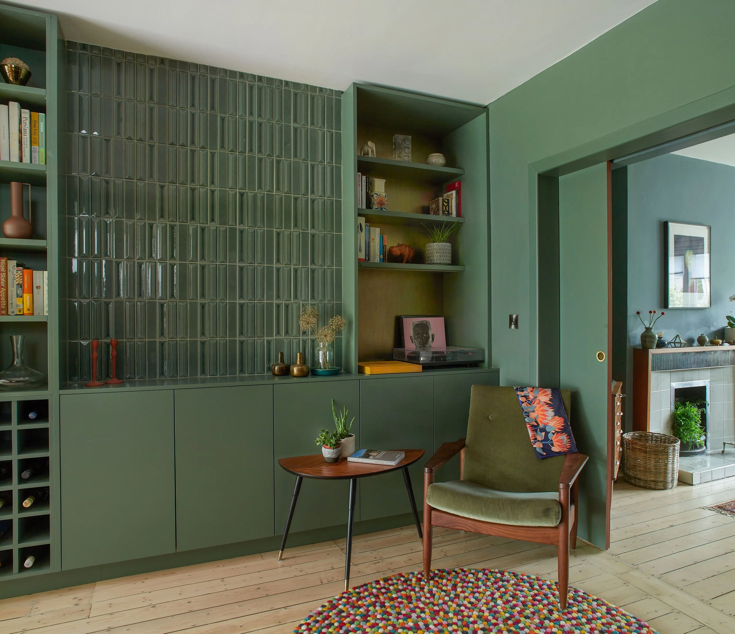A living room with green walls and built-in green cabinets. There is a small wooden table with a potted plant and a magazine, green armchair with a colorful cushion, and a multicolored round rug. A doorway leads to another room with a fireplace and d