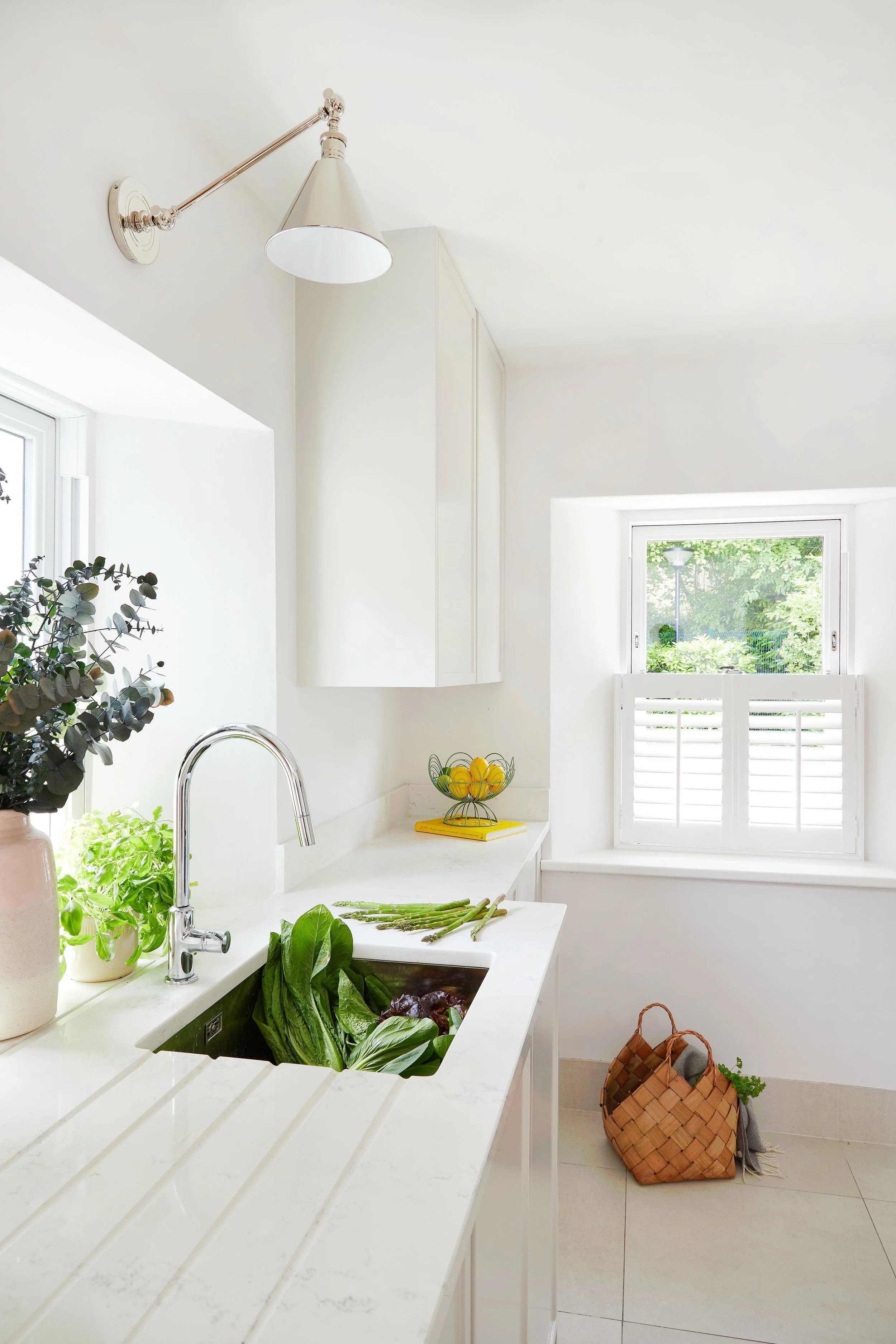Bright white kitchen with a window, a basket of lemons, herbs, and vegetables on the counter, and a woven basket on the floor.