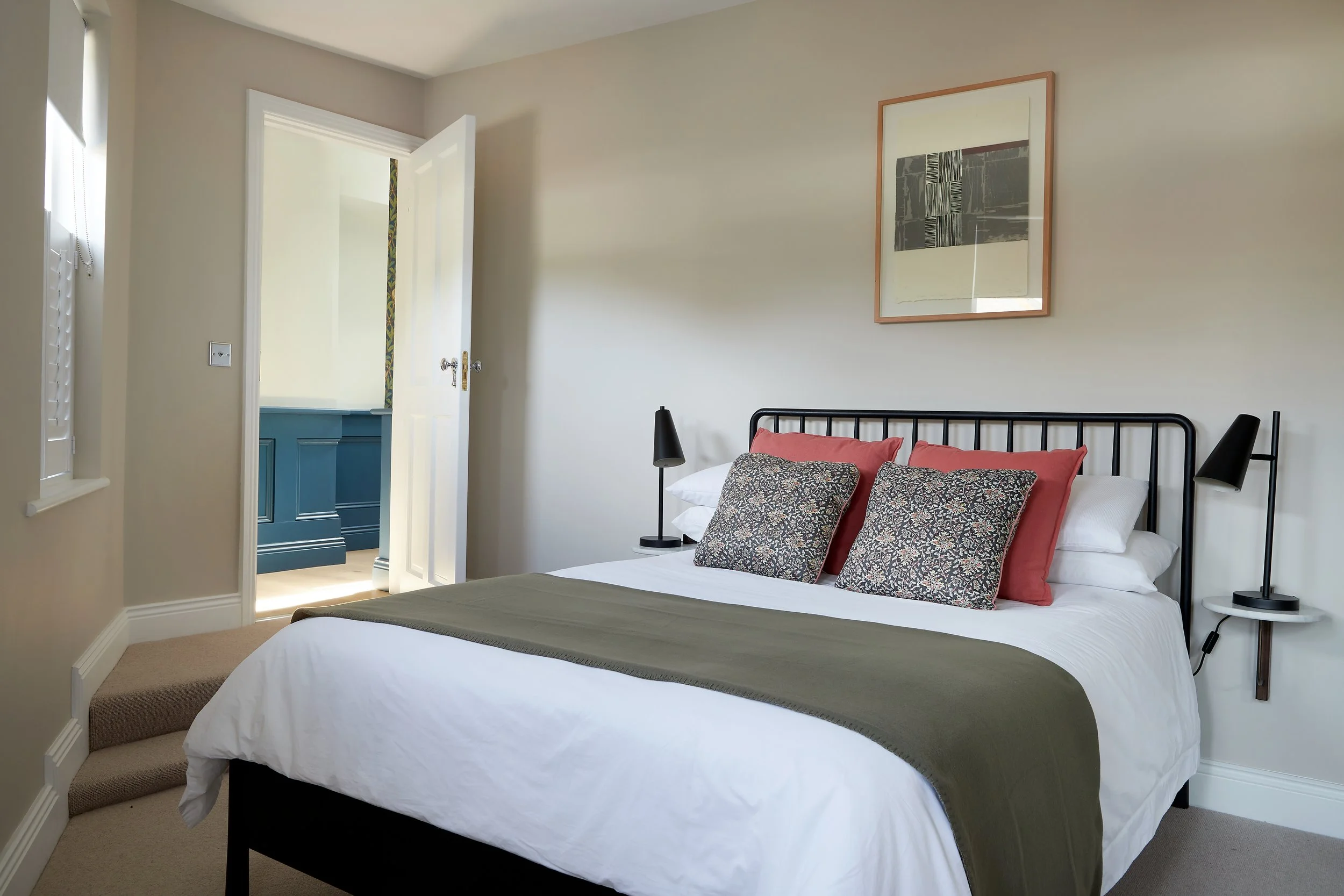 A neatly made bed with white sheets, a green blanket, and decorative pillows in a well-lit bedroom. There are two black wall-mounted lamps on either side of the bed, a framed abstract artwork above the headboard, and an open door leading to another r