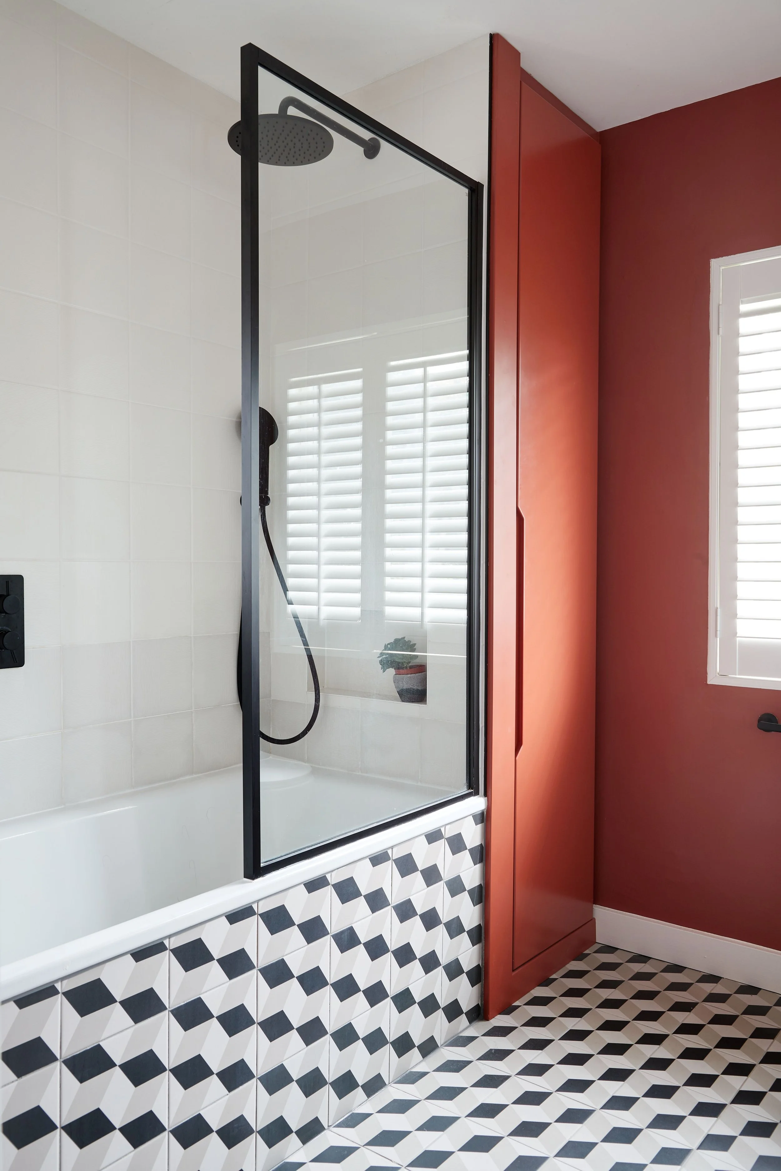 Modern bathroom with a black-framed glass shower door, white tiled walls, patterned floor tiles, a window with white shutters, a terra cotta-colored wall with a built-in cabinet, and a small potted plant on the windowsill.