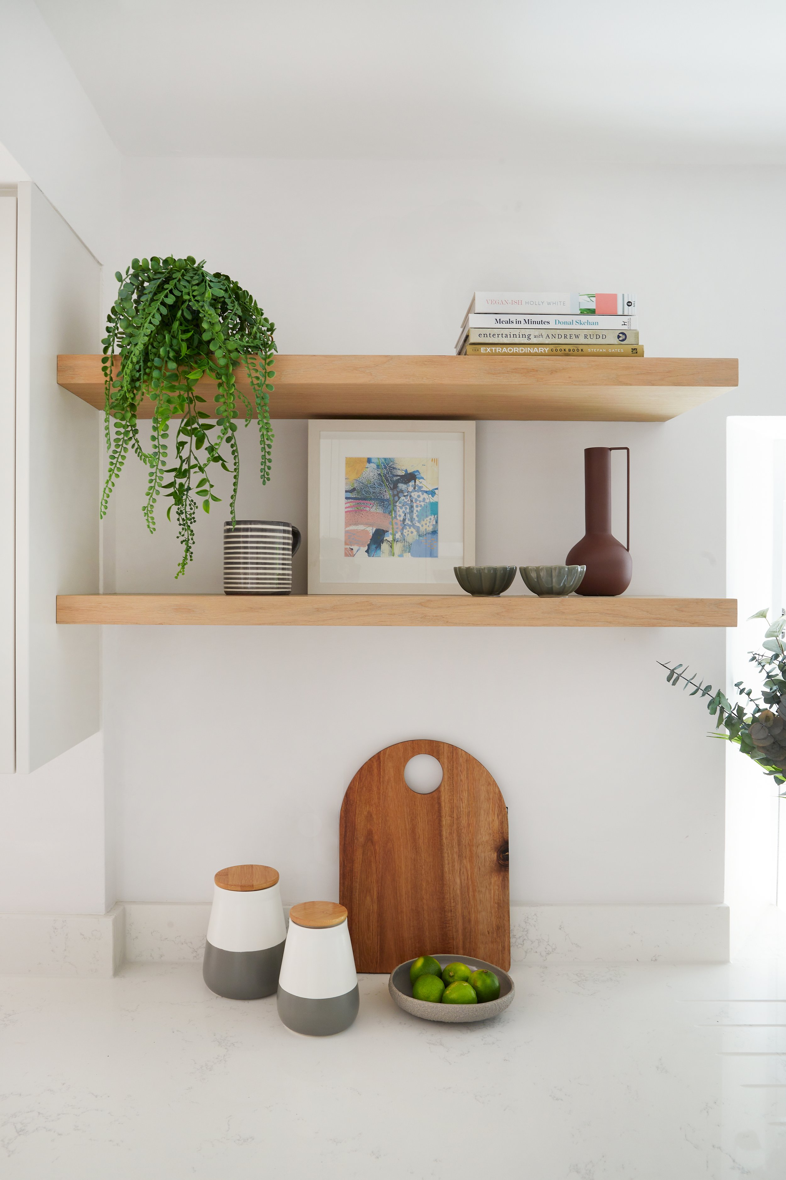 Minimalist kitchen shelf with plants, books, a framed artwork, bowls, a vase, a cutting board, and limes in a dish.