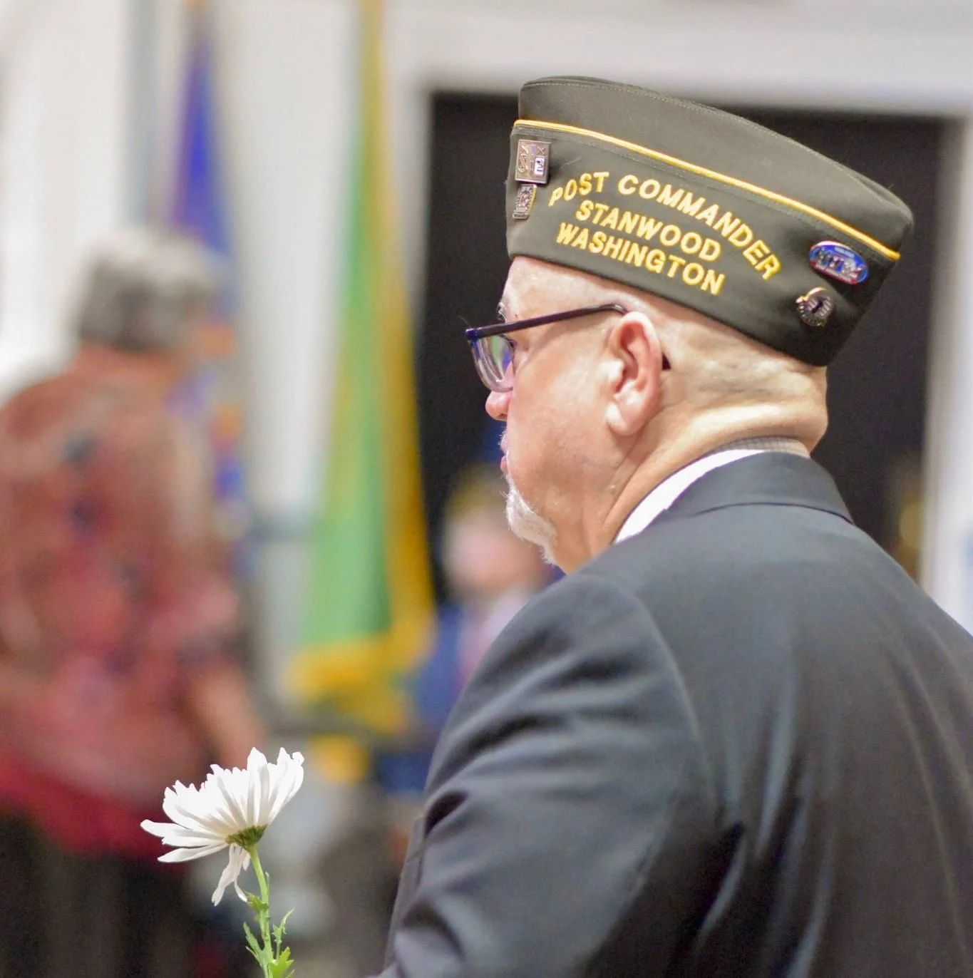 A man wearing a Post Commander hat with name tags stands with a flower in front of him at a formal event.
