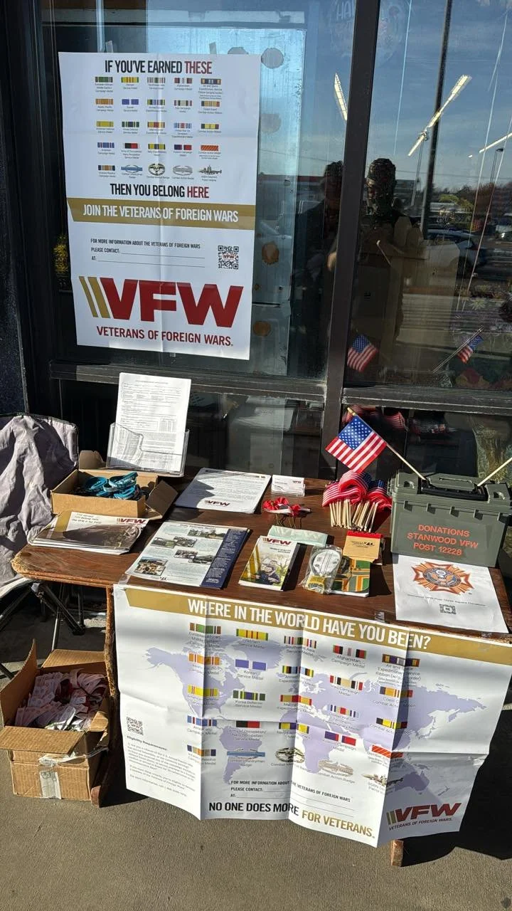 Table set up with pamphlets, American flags, and a donation box outside a building with posters about veterans of foreign wars.