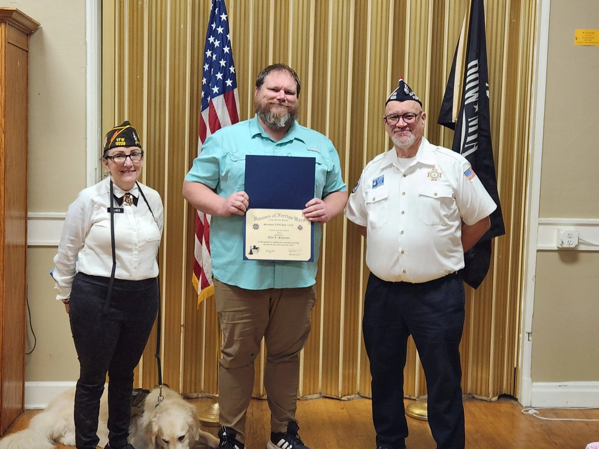Three people posed for a photo, with a woman on the left wearing glasses, a white shirt, and a black hat with a yellow emblem, standing next to a service dog. In the center, a man with a beard holds a certificate, wearing a turquoise shirt. On the right, a man in a white shirt with medals and a black hat with a badge, stands with his hands behind his back. They are in front of American flags and a wooden backdrop.