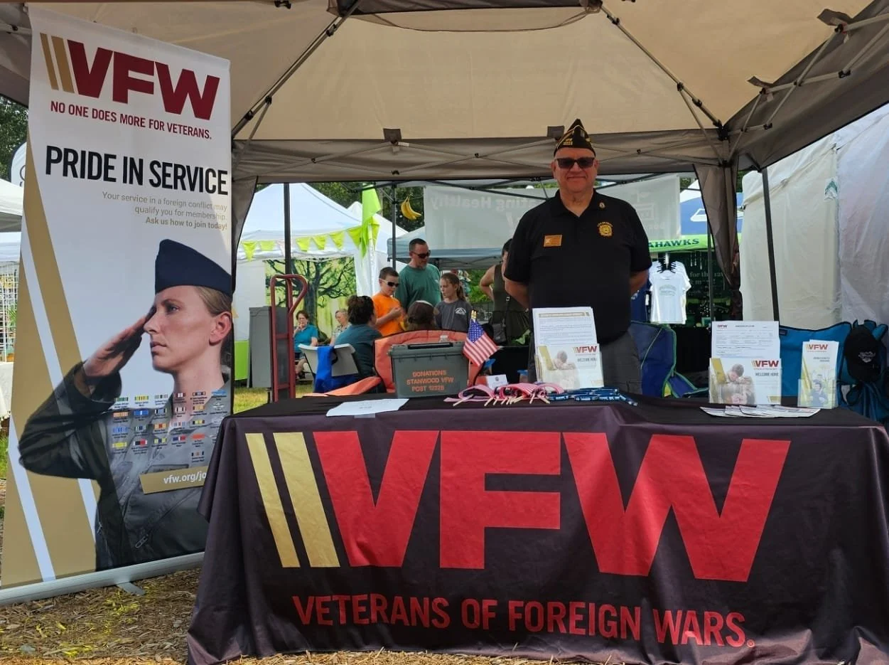 A man in a black Veterans of Foreign Wars uniform standing behind a table at an outdoor event. The table has a black tablecloth with red lettering, displaying the VFW logo and the words "Veterans of Foreign Wars." The table is covered with informational pamphlets, small American flags, and fundraising materials. To the left, there is a large vertical banner with the VFW logo, a saluting woman, and the slogan "No one does more for veterans." In the background, there are other attendees and booths at the event under tents.