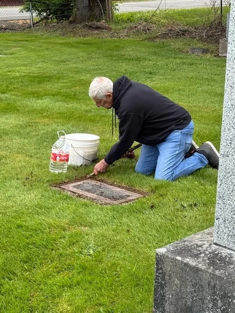 An elderly man with white hair is kneeling on grass, inspecting or cleaning a ground vent or drain cover, with a red-labeled water bottle, a white bucket, and a small white container nearby.