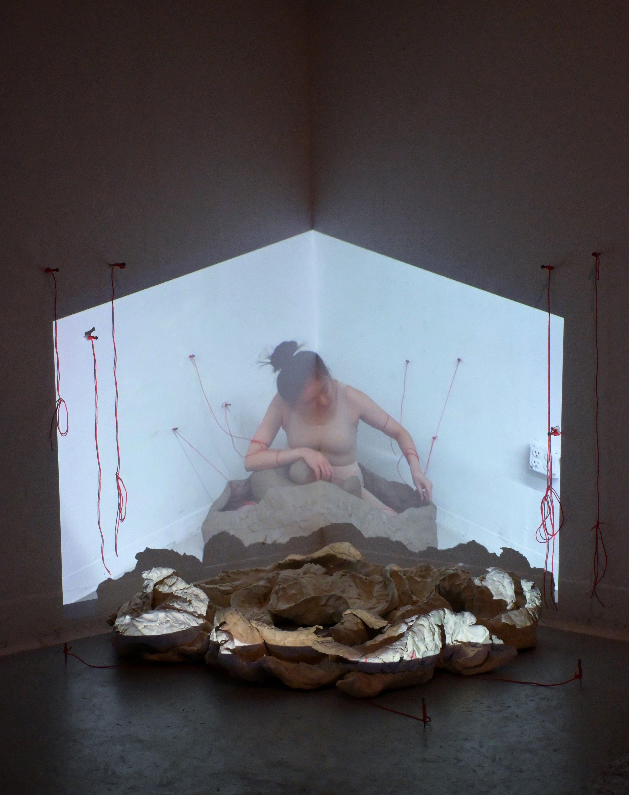 Mixed media art installation depicting a woman emerging from a rocky terrain with wires attached to her arms, a projection of her upper body on the wall, and crumpled paper rocks in front.