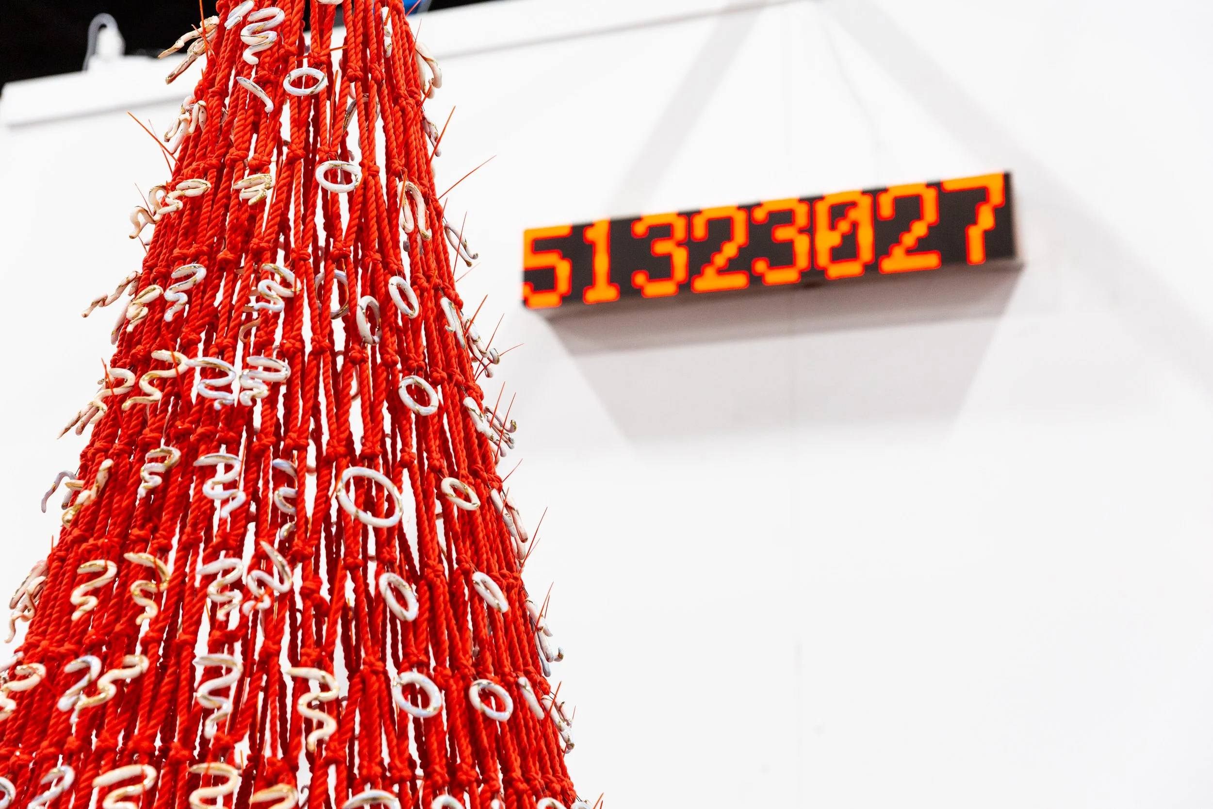 A red and white decorative Christmas tree made of ropes with hooks, hanging in front of a white wall. In the background, a digital display shows the numbers 513223027 in red against a black background.
