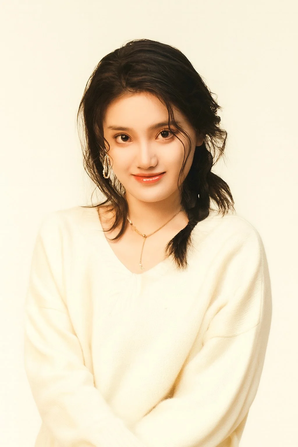 A young woman with dark, wavy hair and a braid over her shoulder, smiling at the camera, wearing a cream-colored sweater and a gold necklace, against a plain light background.