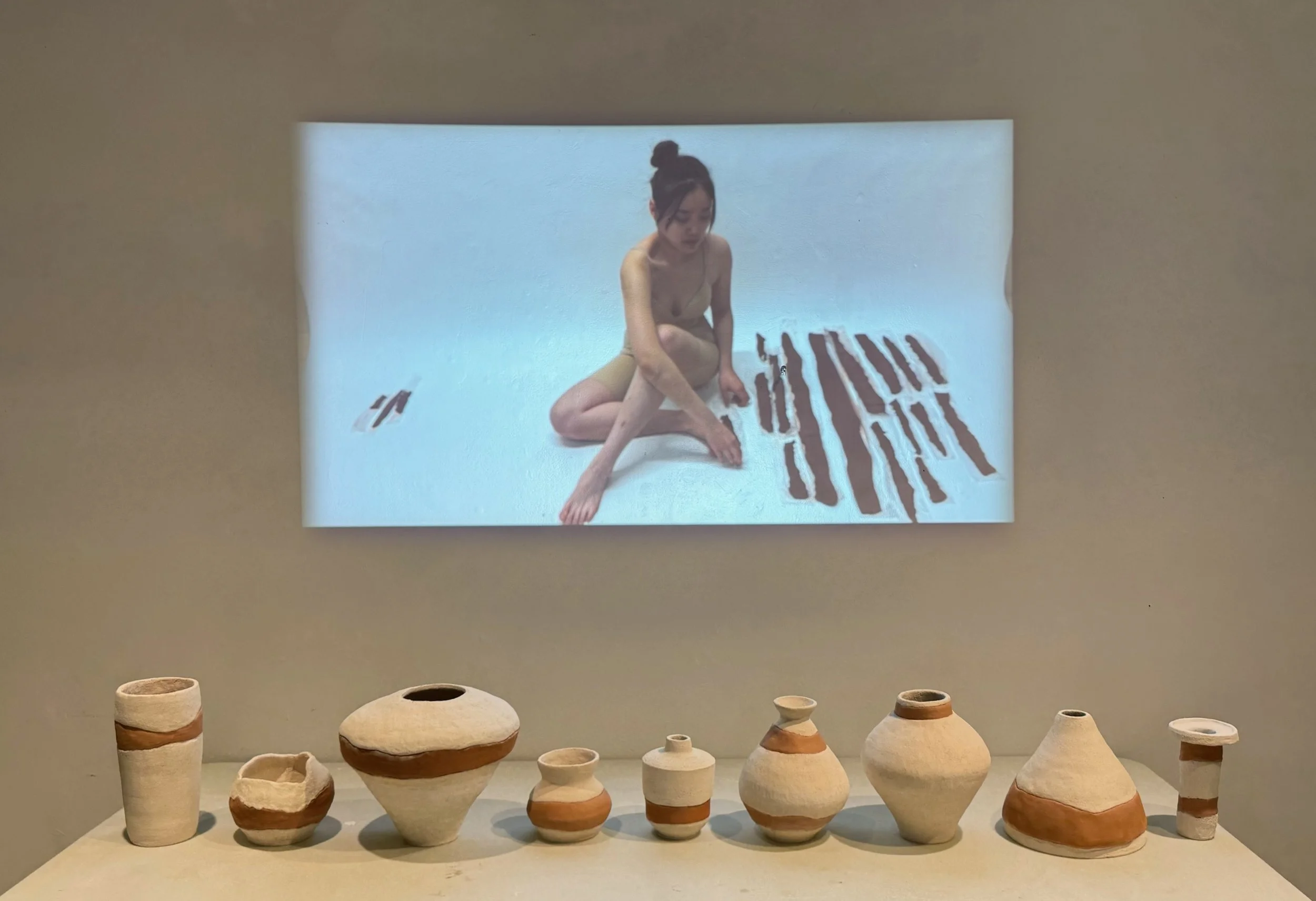 A woman sitting on the ground with folded legs in front of a light background, demonstrating a piece of art with black vertical lines. On the table below are nine ceramic pots of various shapes, sizes, and earth-tone colors.