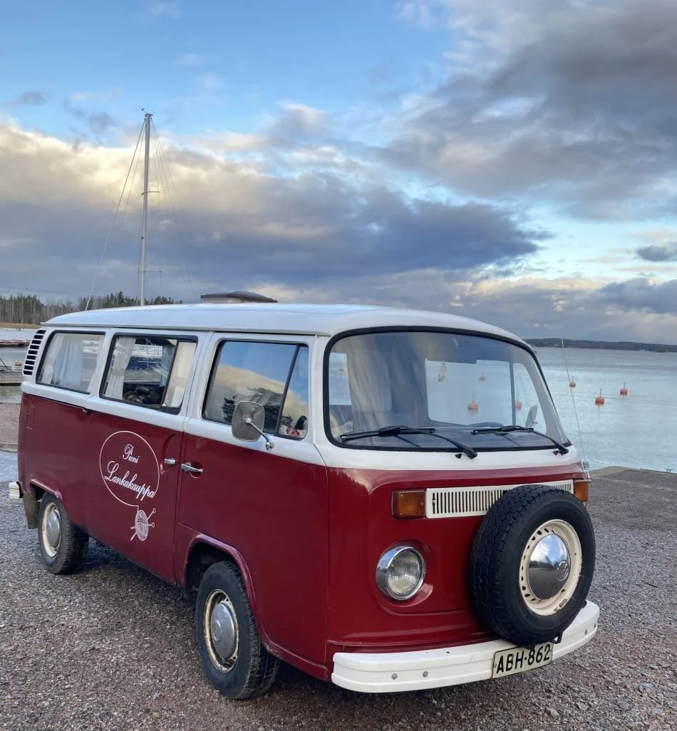 Punainen ja valkoinen vanha Volkswagen Transporter - pysäköity meren rannalle, taivaalla pilviä ja veneitä vedessä.