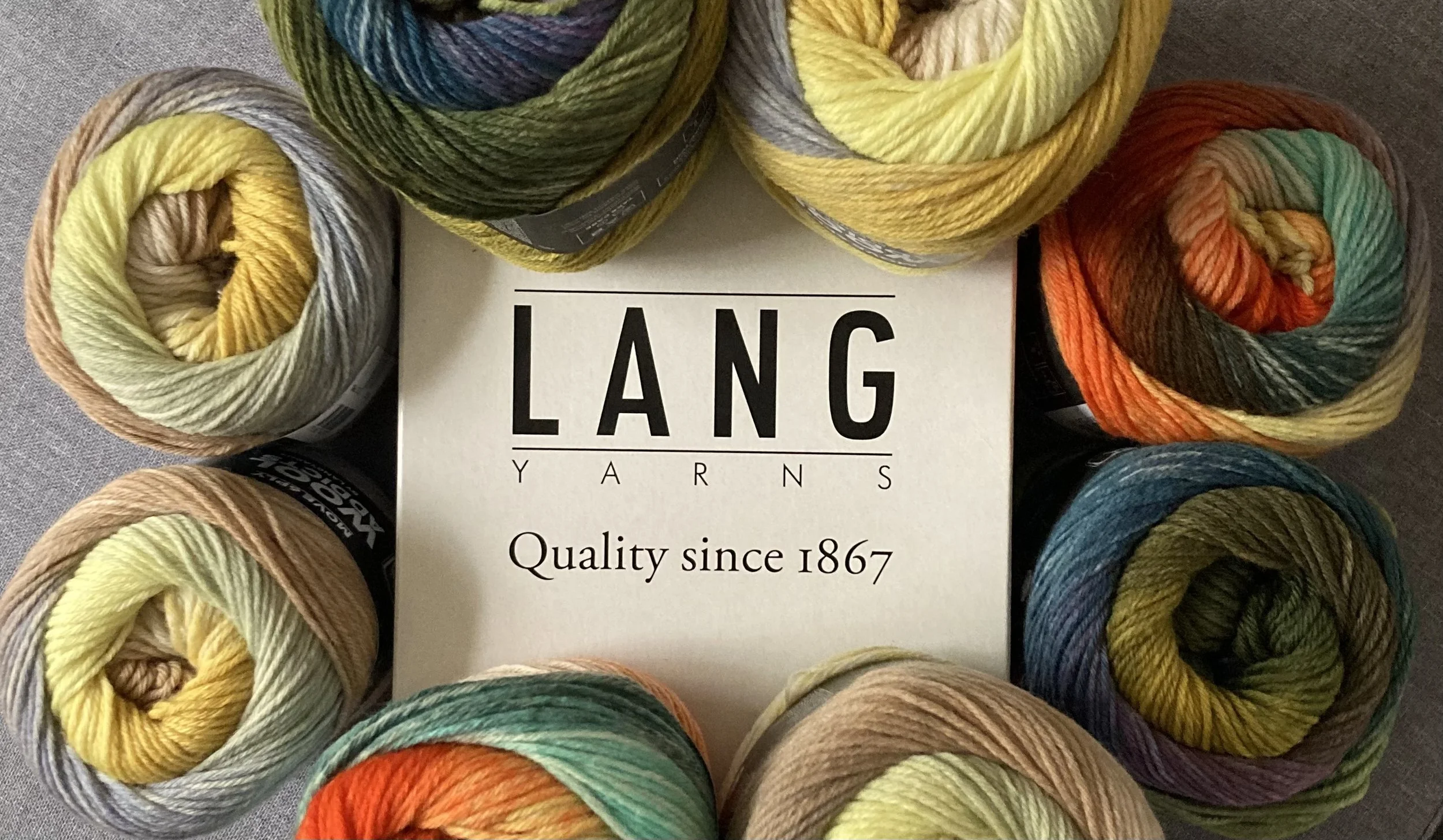 Lang Yarns -lankoja, luonnonvärisiä ja kirkkaita sävyjä, asetettuna ympärilleen spiraalimaisiksi rulliksi vierekkäin, ja keskellä valkoinen kortti.