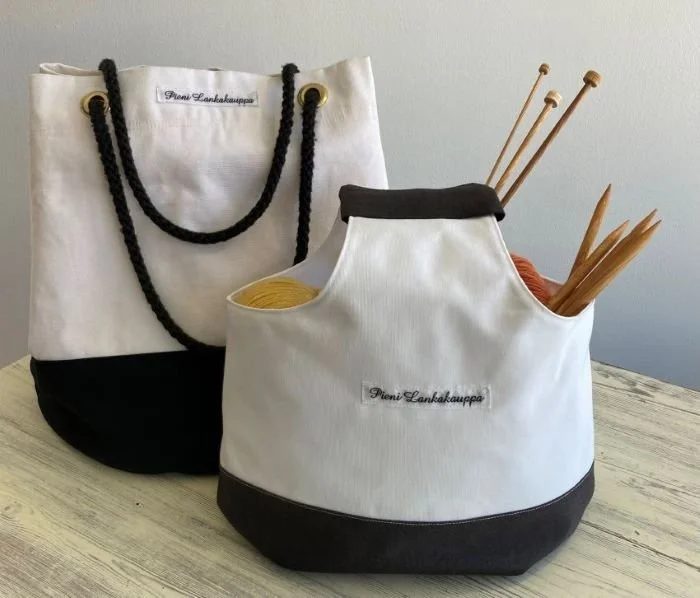 Kaksi valkoista ja mustaa kangaspussia, joiden sisällä on neulepuikkoja, pöydän pinnalla.