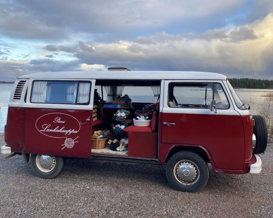 Punainen ja valkoinen vanha Volkswagen pakuauto, joka on muokattu liikkuvaksi lankakaupaksi. Autossa on myyntitiski ja myytävänä erilaisia lankoja ja muita tuotteita.