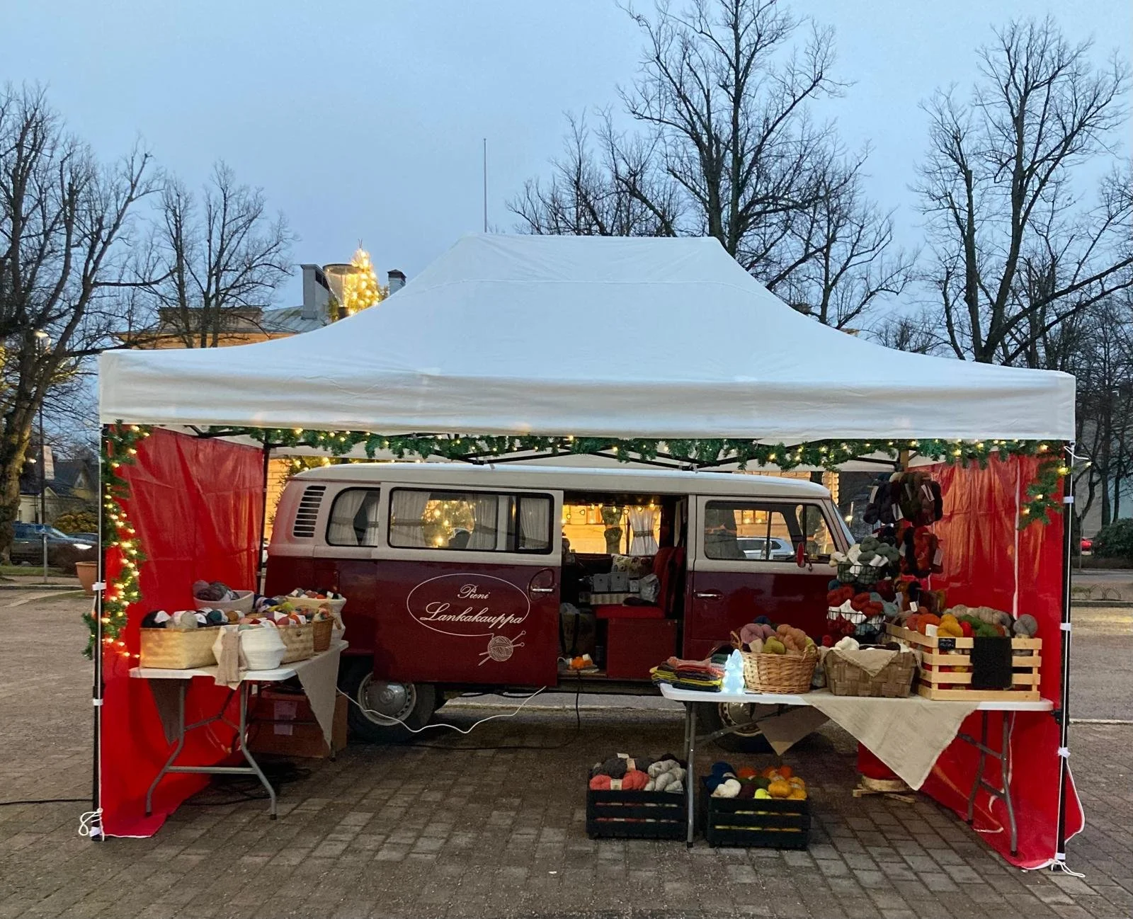 Kuvassa on joulumyyntikoju, jossa myydään lankoja ja käsitöitä. Kojun takana on auto ja ympärillä on joulukoristeita ja valoja. Taustalla on taloja ja talvimaisema.