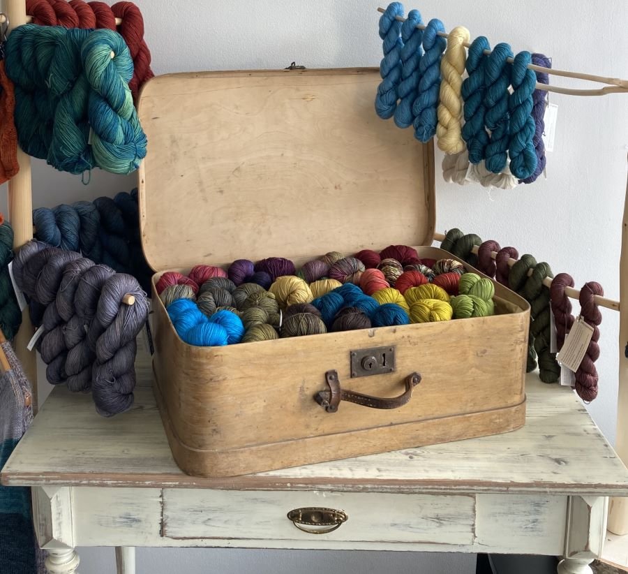 Vanha matkalaukku sisältää monivärisiä lankakeriä, joiden ympärillä roikkuu myös lankoja. Lankakerät ovat eri väreissä, kuten sinistä, punaista, keltaista ja vihreää.