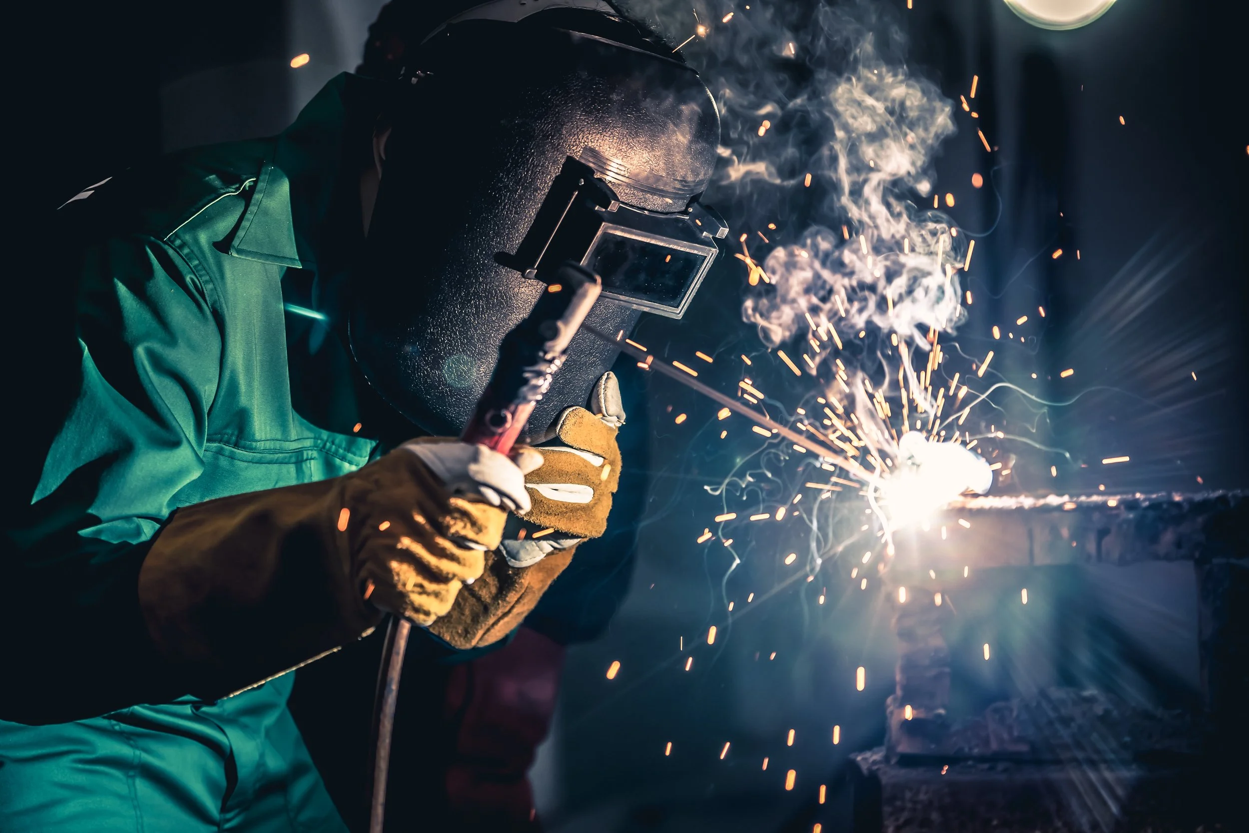 A person welding metal, wearing safety gloves and a welding mask, with sparks and smoke flying around in a dark workshop or outdoor setting.