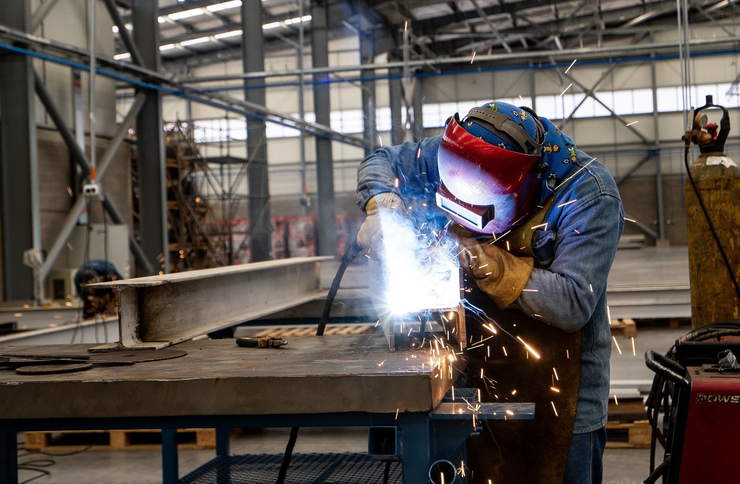 A worker welding metal on an industrial construction site, wearing protective gear including a welding helmet and gloves.