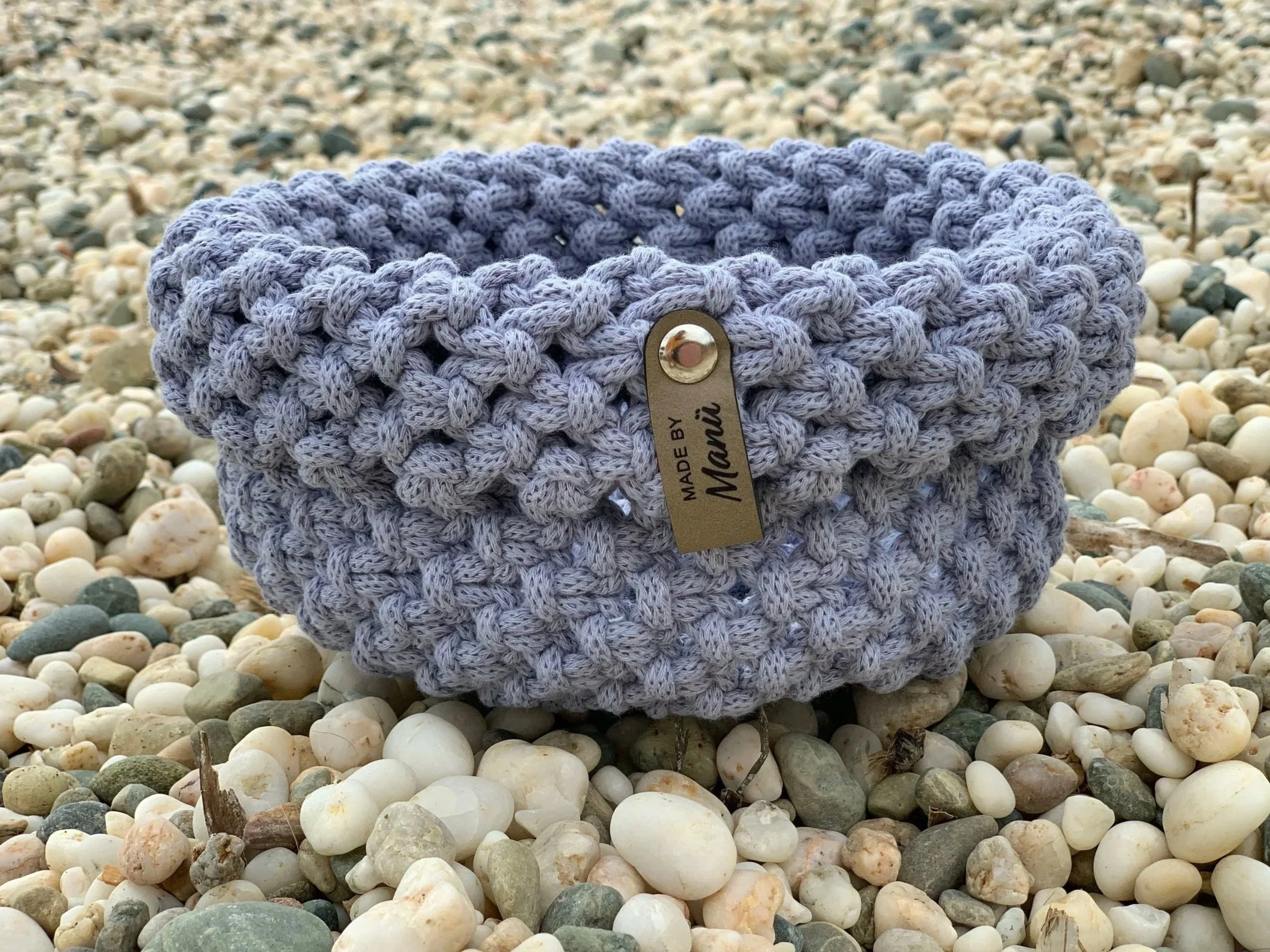 Ein handgefertigter, grau geflochtener Korb mit einer Metallbeschriftung, der auf einem Kieselsteinstrand liegt.