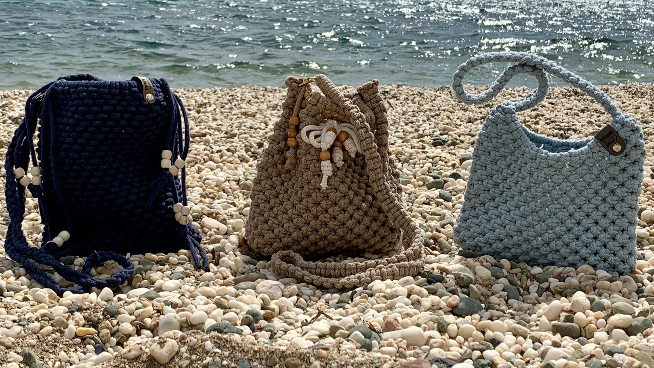 Drei handgefertigte, geknüpfte Taschen in den Farben navyblau, beige und hellblau, stehen auf einem kiesbedeckten Strand vor dem Meer.