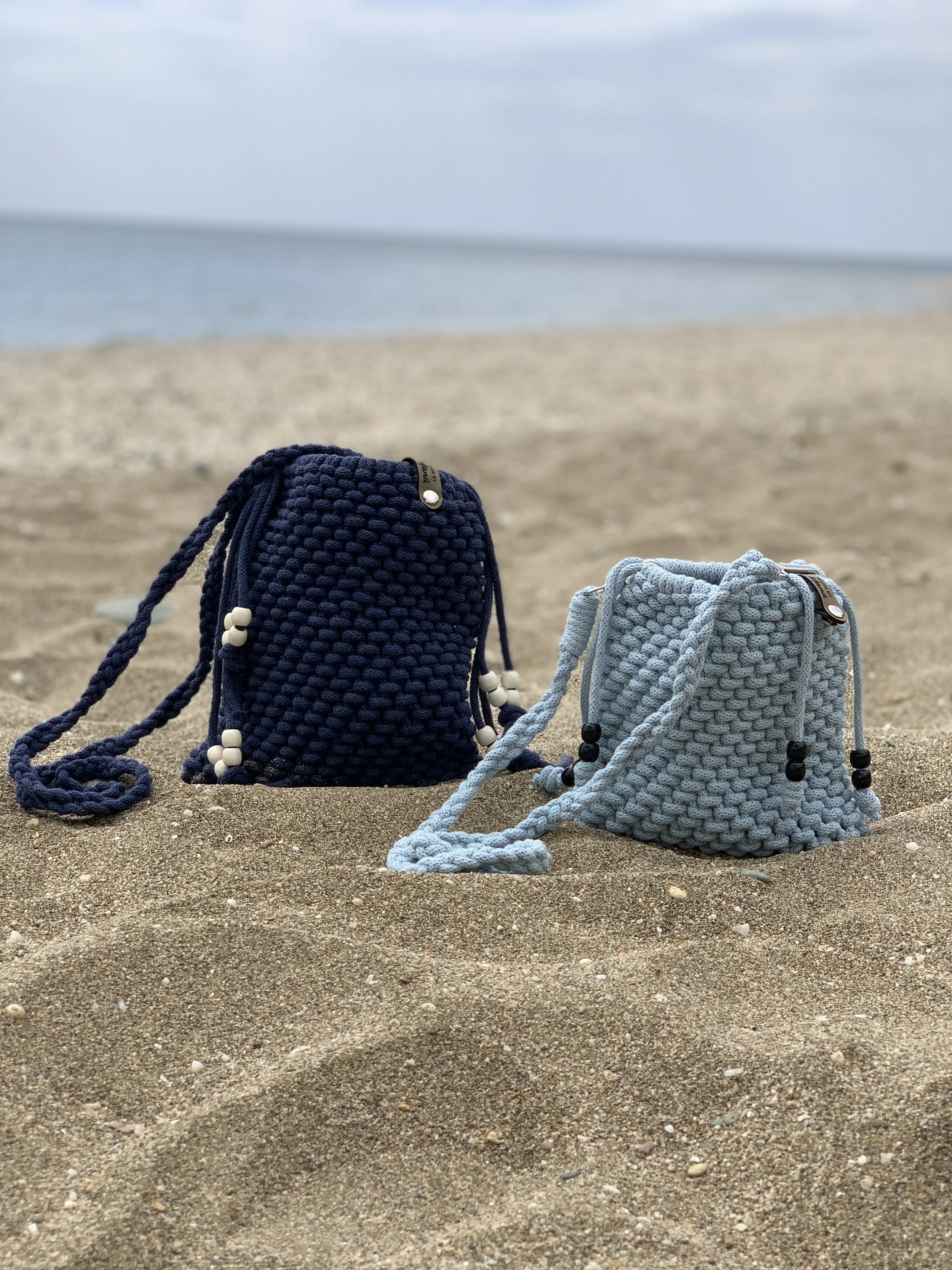 Zwei aufwendig gestrickte Taschen, eine in Dunkelblau und die andere in Hellgrau, stehen auf einem Sandstrand. Im Hintergrund ist das Meer und ein bewölkter Himmel sichtbar.