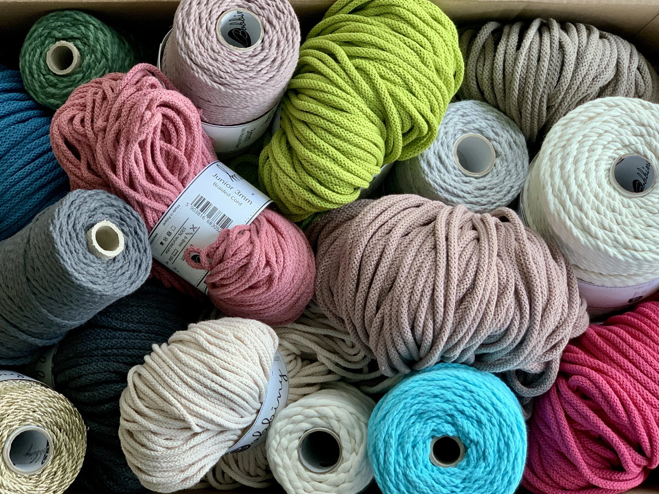 Bunte Knäuel aus Garn in verschiedenen Farben wie rosa, grün, blau, beige, grau, weiß und pink, liegen in einer Kiste.