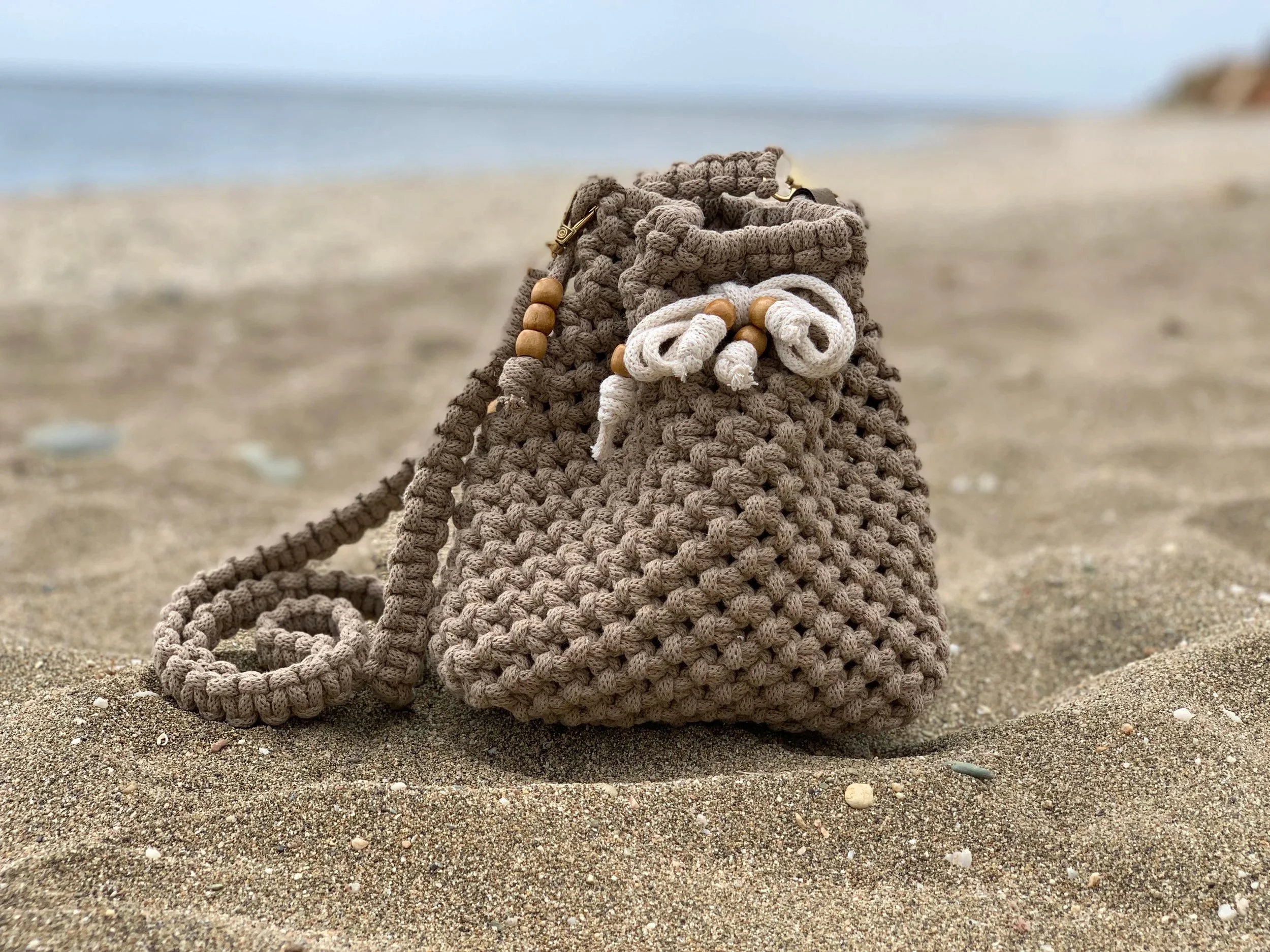 Ein aus geflochtenem Seil bestehender Beutel mit Holzperlen auf einem Sandstrand, im Hintergrund das Meer und ein Himmel.