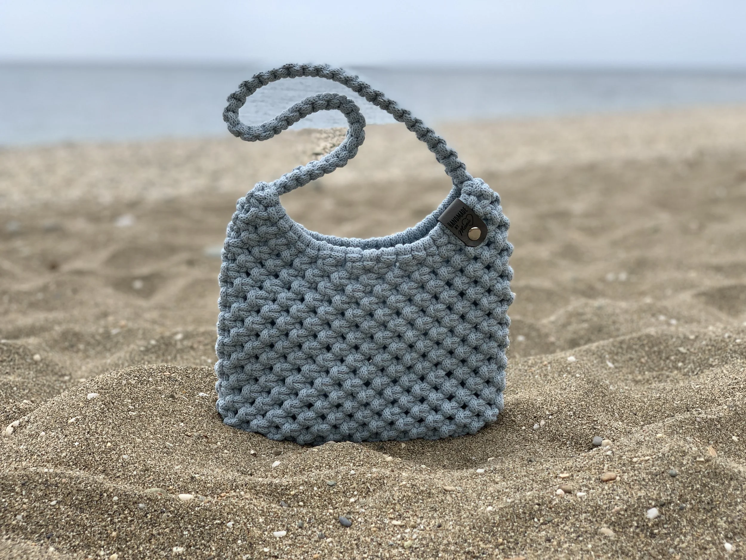 Ein gestrickter Beutel auf einem Sandstrand mit Blick auf den Ozean im Hintergrund.