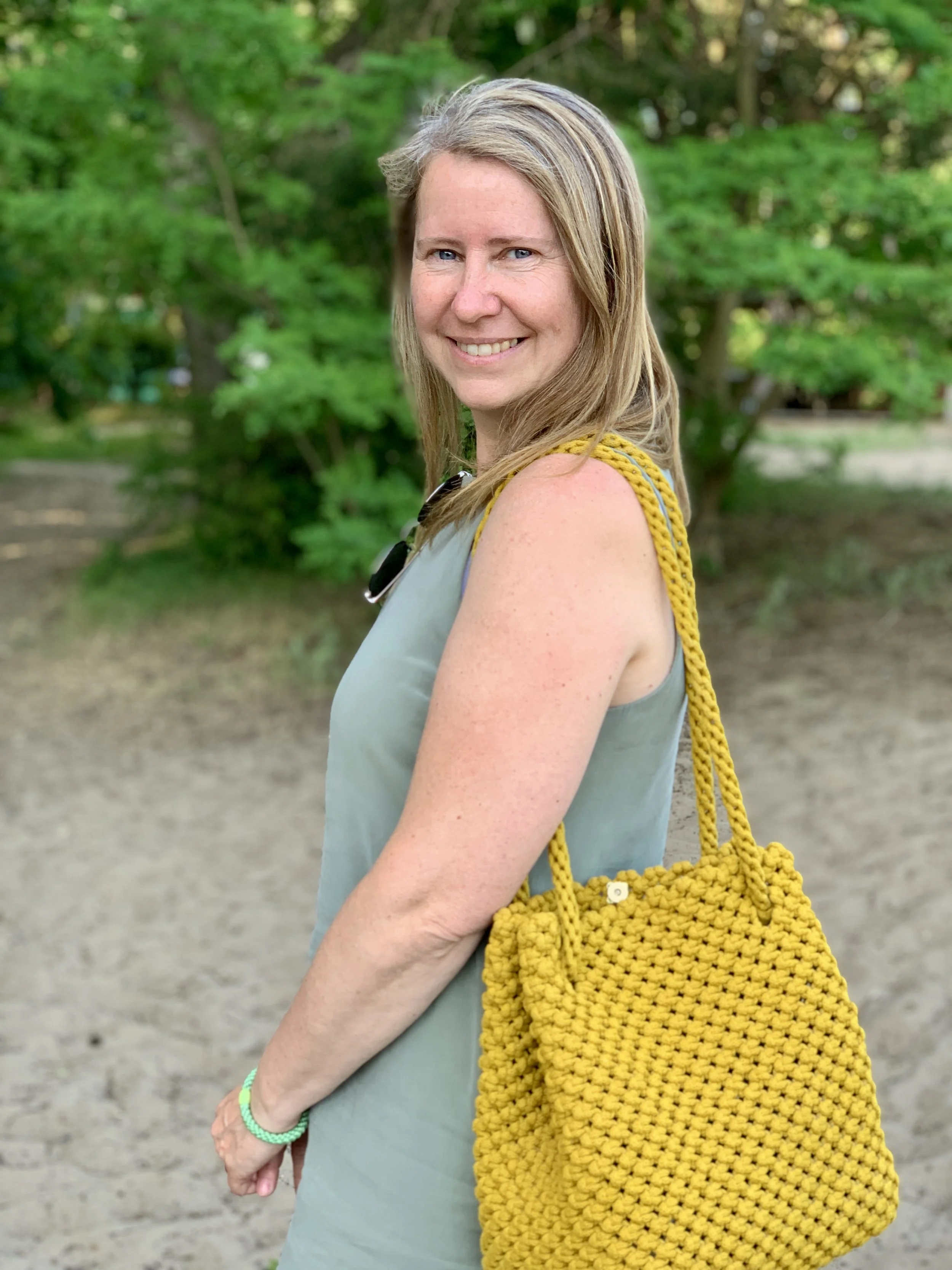 Porträt einer lächelnden Frau mit braunen Haaren, hellblauen Augen, in einem grünen Kleid und einer gelben, handgestrickten Tasche, im Freien mit grünen Bäumen im Hintergrund.
