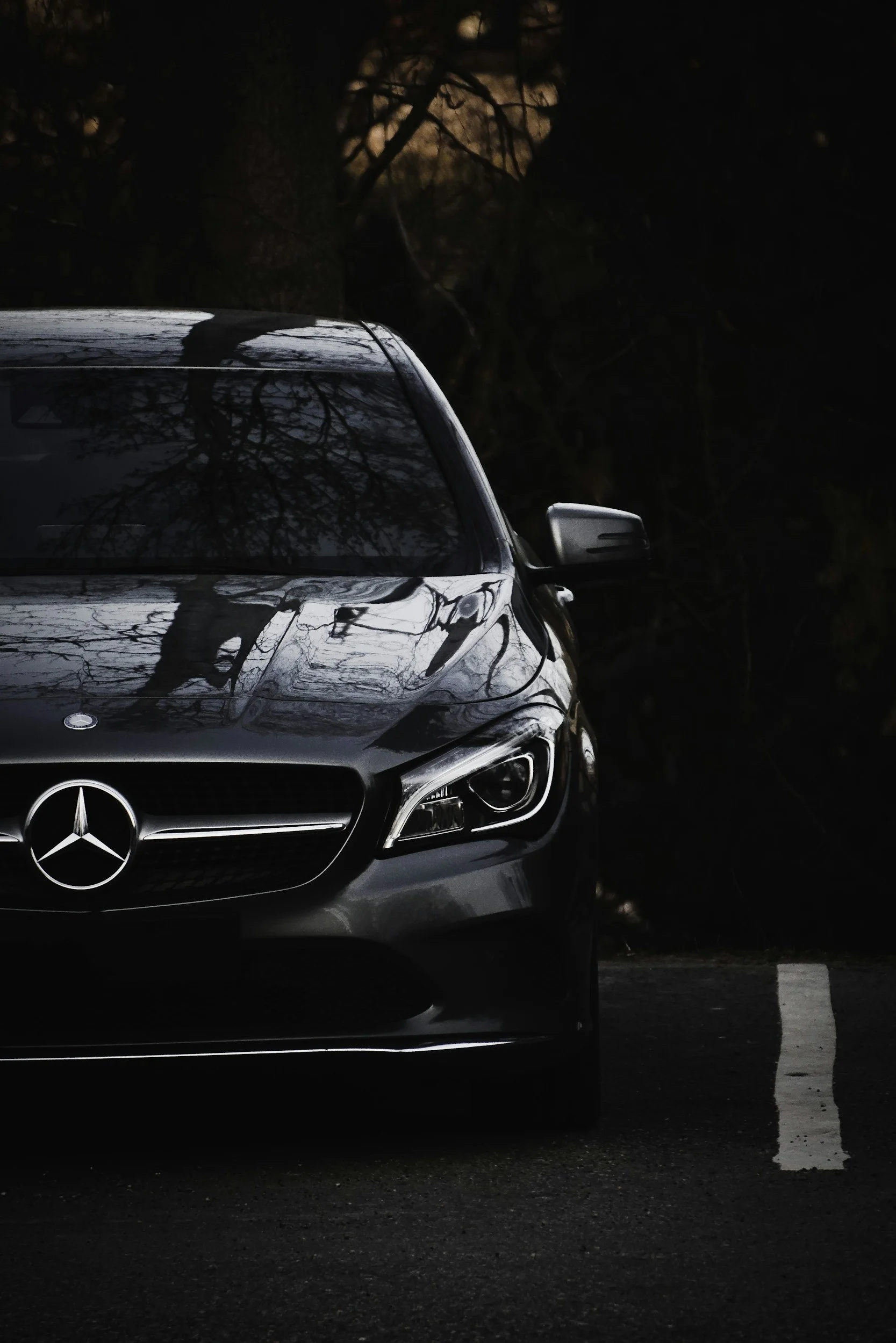 Voiture Mercedes noire garée dans une forêt sombre, reflet des arbres sur la carrosserie, prise de vue en gros plan à l'avant