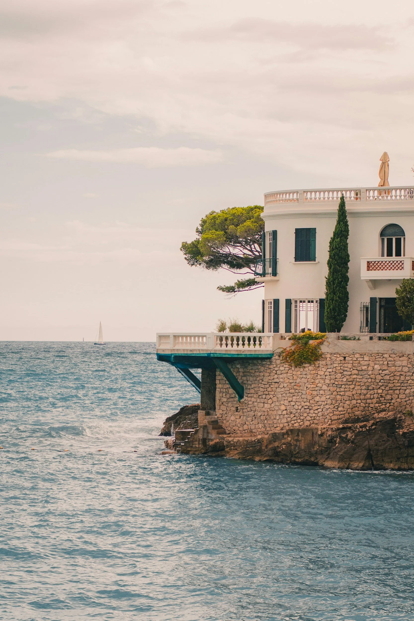 Maison blanche en bord de mer avec une terrasse surplombant l'eau, un arbre et un cactus devant, ciel nuageux.