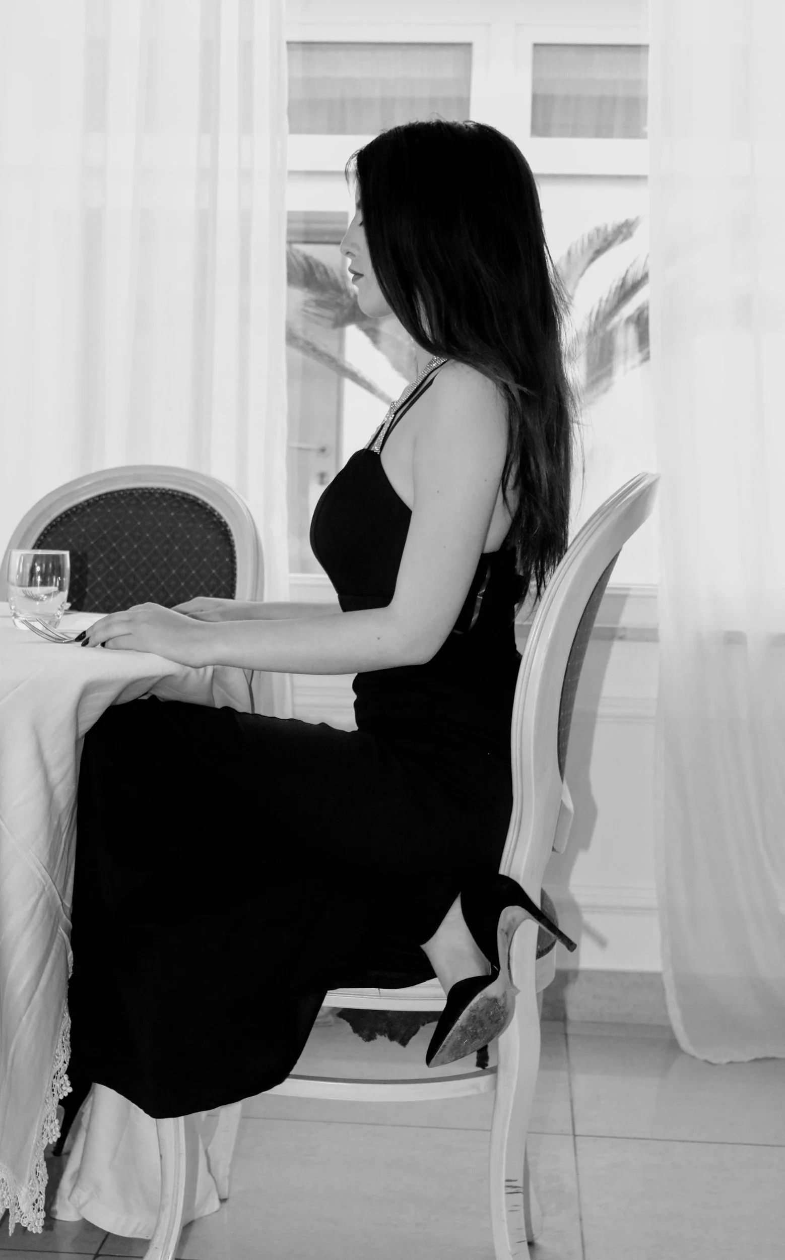 Une femme en robe noire assise à une table, vue de profil, dans une ambiance élégante, avec des draperies blanches en arrière-plan.