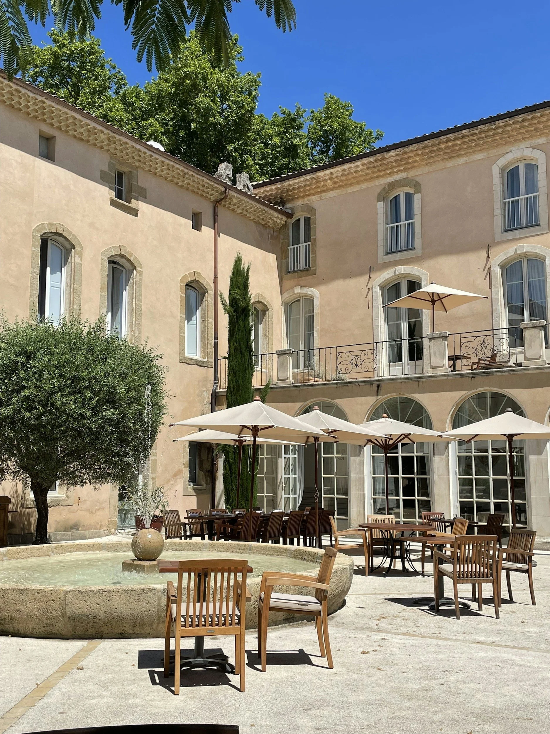 Cour intérieure d'un bâtiment résidentiel avec une terrasse, des tables et chaises en bois, des parasols et un bassin avec une fontaine.
