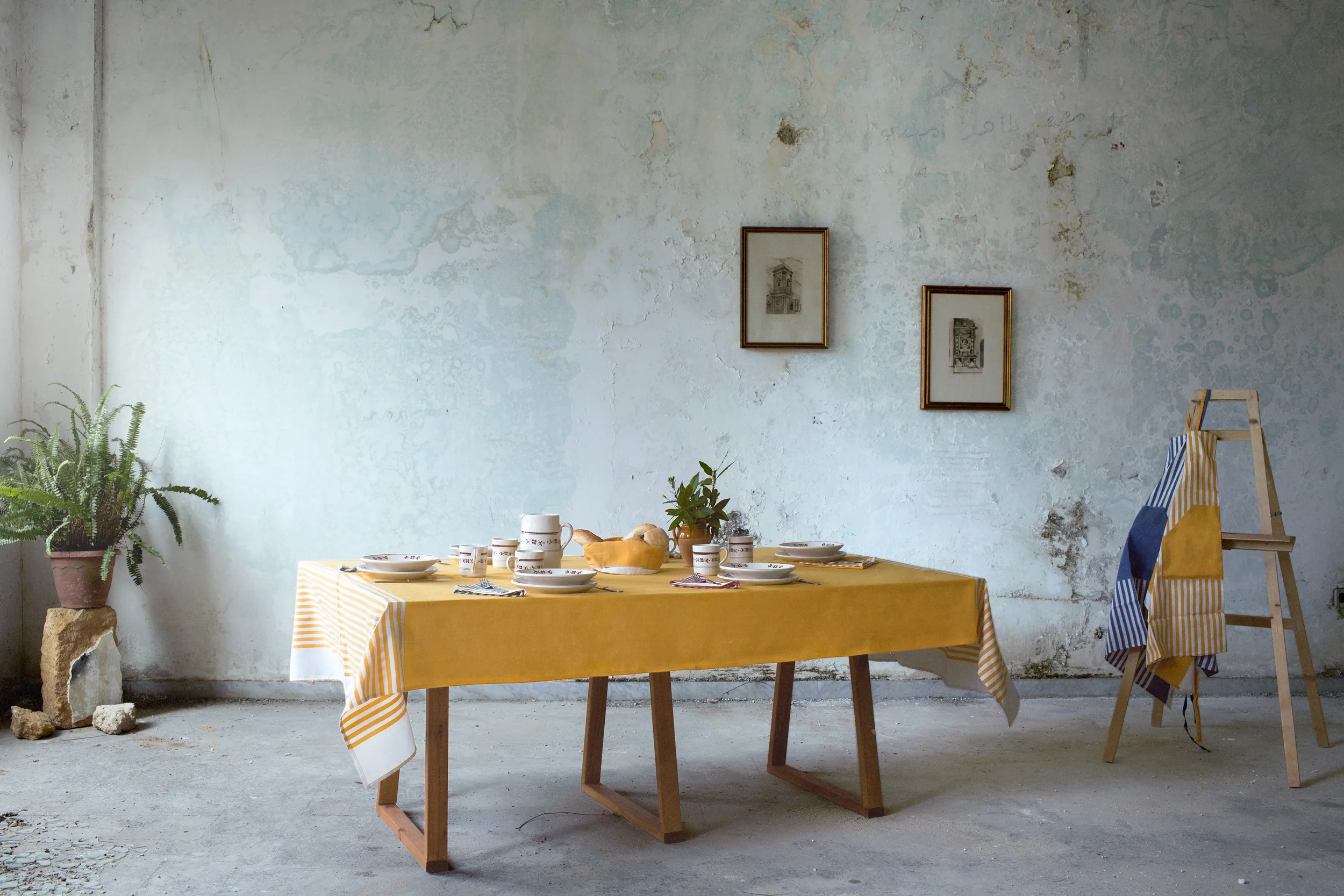 A rustic dining room with a yellow tablecloth-covered table set with plates, cups, and a basket of bread, against a distressed white wall with two framed sketches and a potted plant nearby.