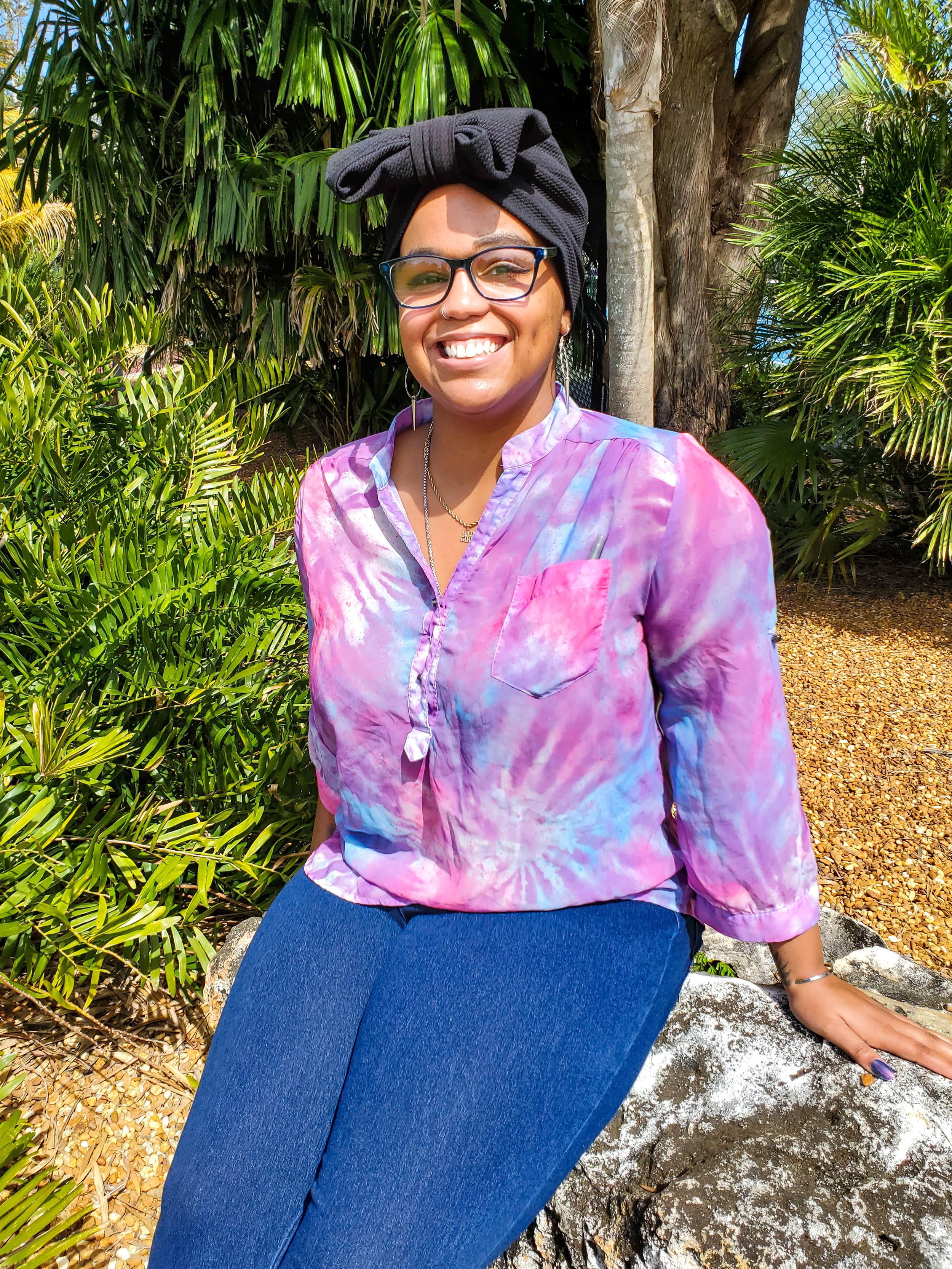 A woman smiling and sitting outdoors near lush green foliage and trees, wearing glasses, a black head wrap, a colorful pink and purple tie-dye shirt, and blue pants.