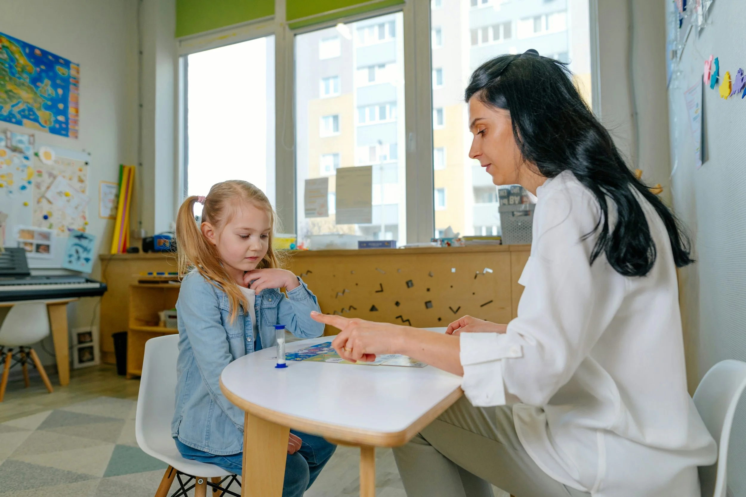 A young girl with red hair sitting at a table with a woman, likely a healthcare professional, in a room with colorful children's artwork and large windows showing apartment buildings outside.