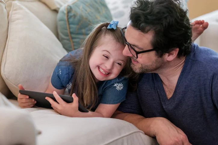 A man and a young girl are lying on a couch, laughing and sharing a moment together, with the girl holding a tablet.